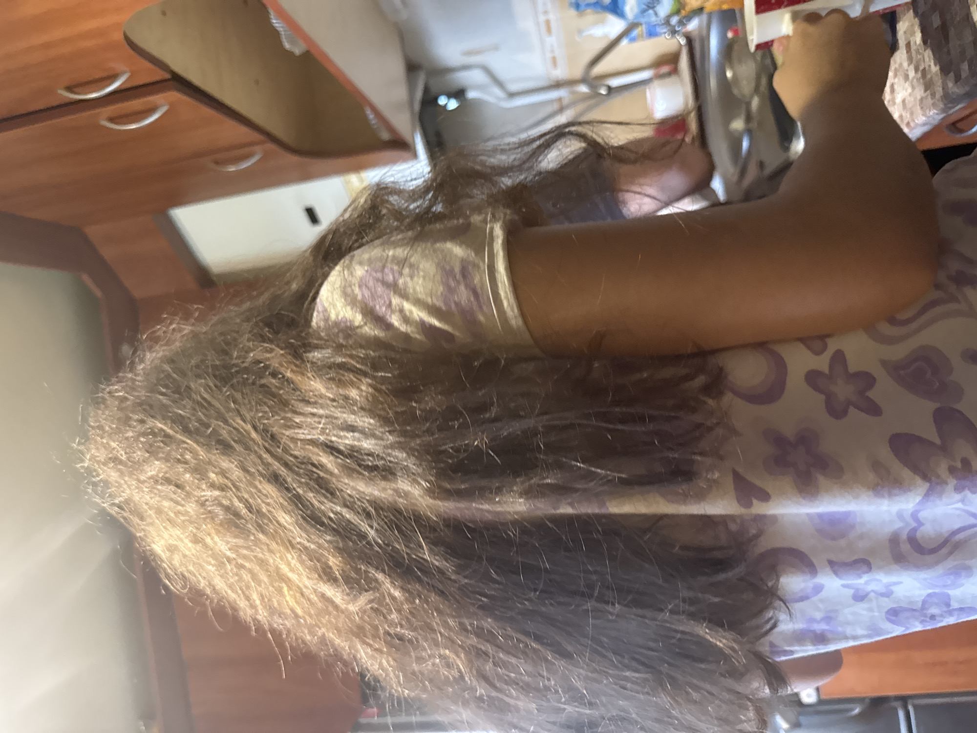 A person with long, brown, slightly disheveled hair is shown from behind in a kitchen. The person is wearing a light-colored t-shirt with a purple floral pattern. Their right arm is visible, and they are holding a red and white cup. In the background, wooden cabinets are mounted above a countertop. A sink with a faucet is also visible. The overall lighting suggests an indoor environment during daylight hours.