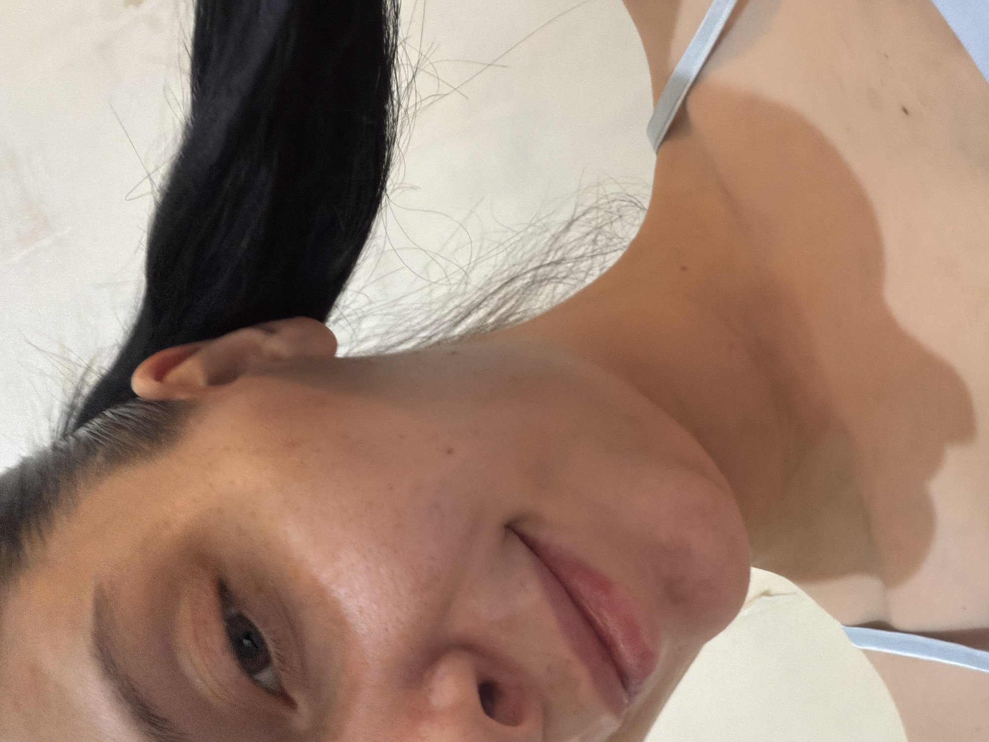 The image shows a close-up, low-angle shot of a young woman's face and upper body. She has dark hair pulled back in a ponytail, and her skin has a light tan with freckles visible on her cheek. Her eye is dark brown, and she has a slight smile. She is wearing a light blue strap, possibly from a tank top or swimsuit. The background is a plain, light-colored wall. The image is a selfie, taken indoors, and there is no discernible information about the location in Tbilisi, Georgia.