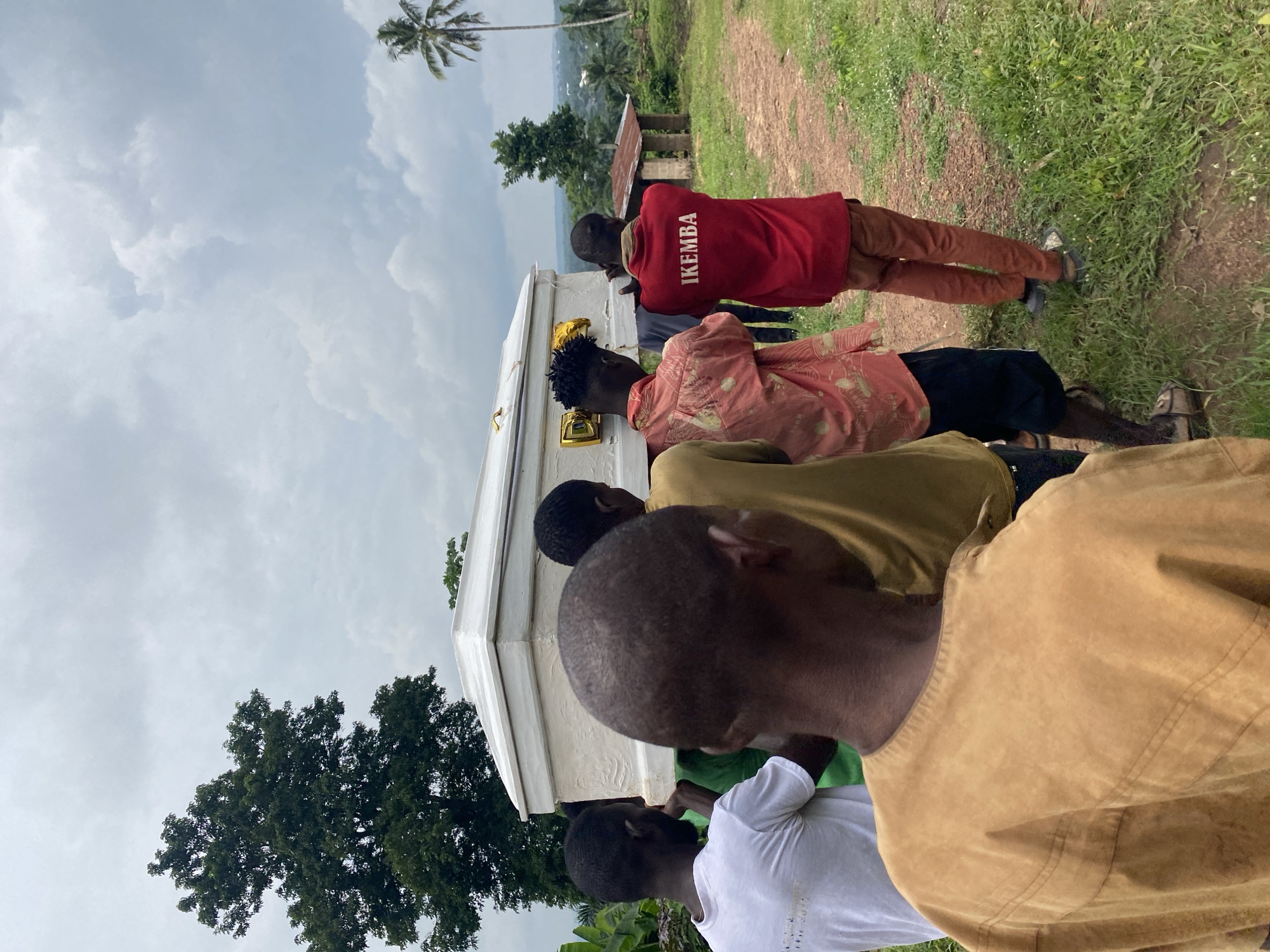 In Achara, Nigeria, a group of men are carrying a white casket through a rural, grassy area. The sky is overcast with grey clouds. A large tree with dark green foliage stands to the left of the frame. In the distance, a small, rustic structure with a tin roof is visible. To the right, a palm tree reaches towards the sky. One of the pallbearers is wearing a red shirt with the word "IKEMBA" printed on the back in white letters. The casket is adorned with golden accents. The ground is a dirt path covered with grass.