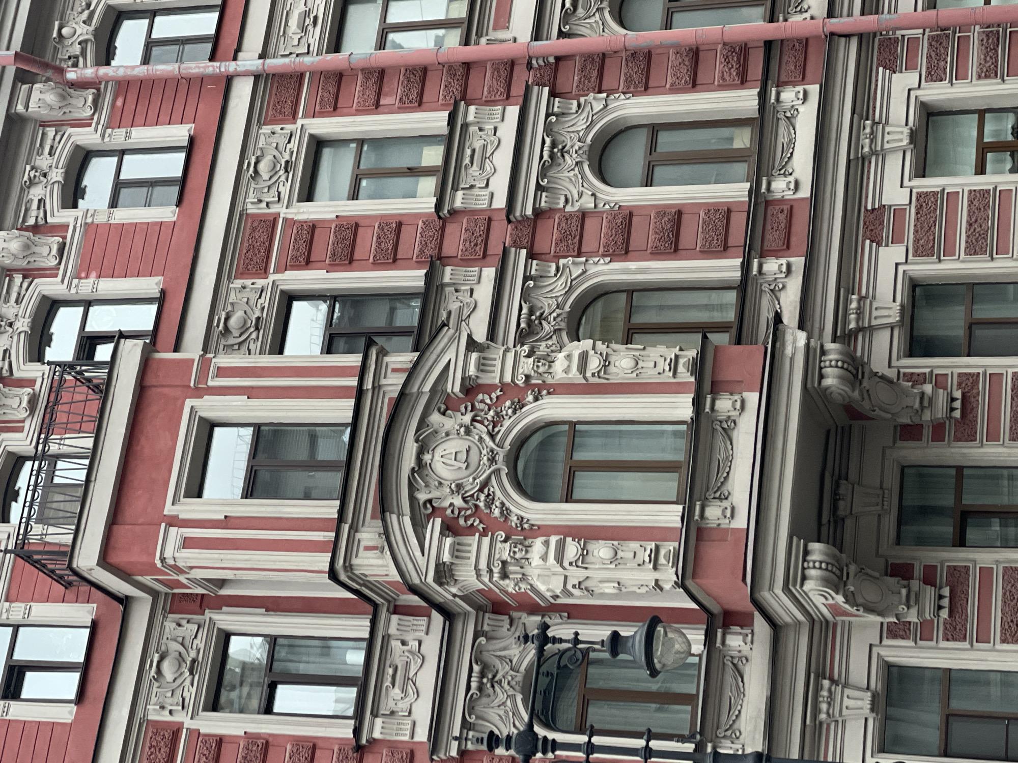 The image shows the facade of a building in Saint Petersburg, Russia, characterized by its elaborate architectural details. The building features a predominantly red and white color scheme. The windows are rectangular and arched, each surrounded by ornate, sculpted frames. Decorative elements such as friezes, molding, and other architectural embellishments are visible throughout the facade. A balcony with wrought iron railings is present on the left side of the image. A street lamp is seen towards the lower part of the image. A pipe is on the top part of the image.