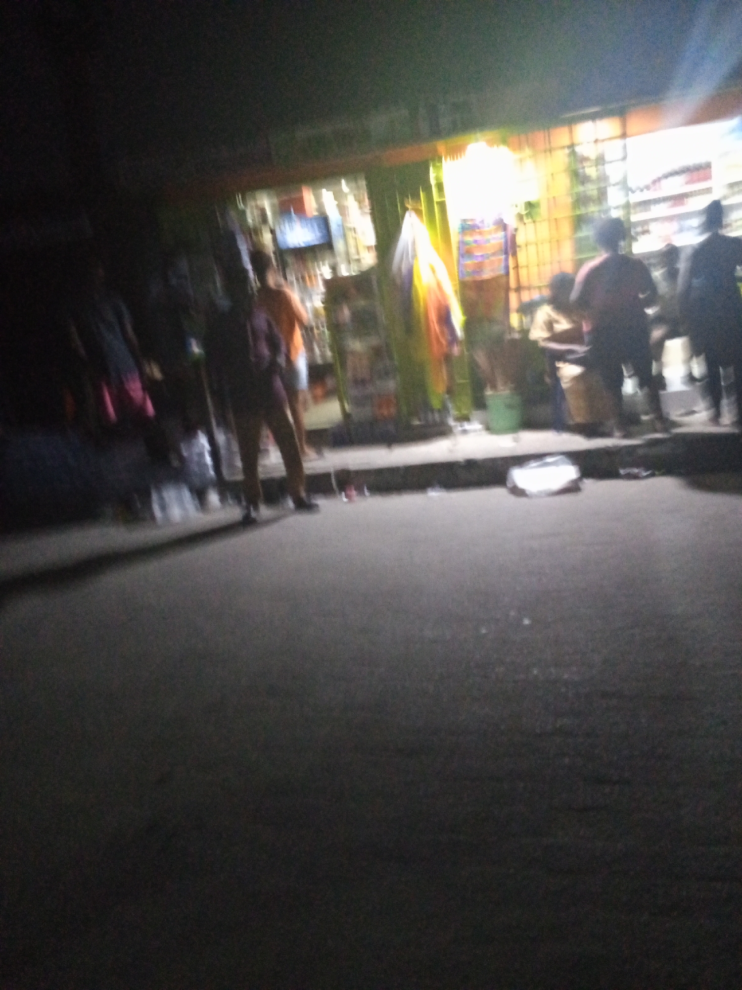 The image depicts a nighttime scene in Ado, Nigeria, outside a brightly lit storefront. Several people are gathered in front of the store, some standing and others sitting. The store has an open front, displaying merchandise, and possibly a television inside. The road in front of the store is dark, and a discarded piece of white material lies on the ground.
