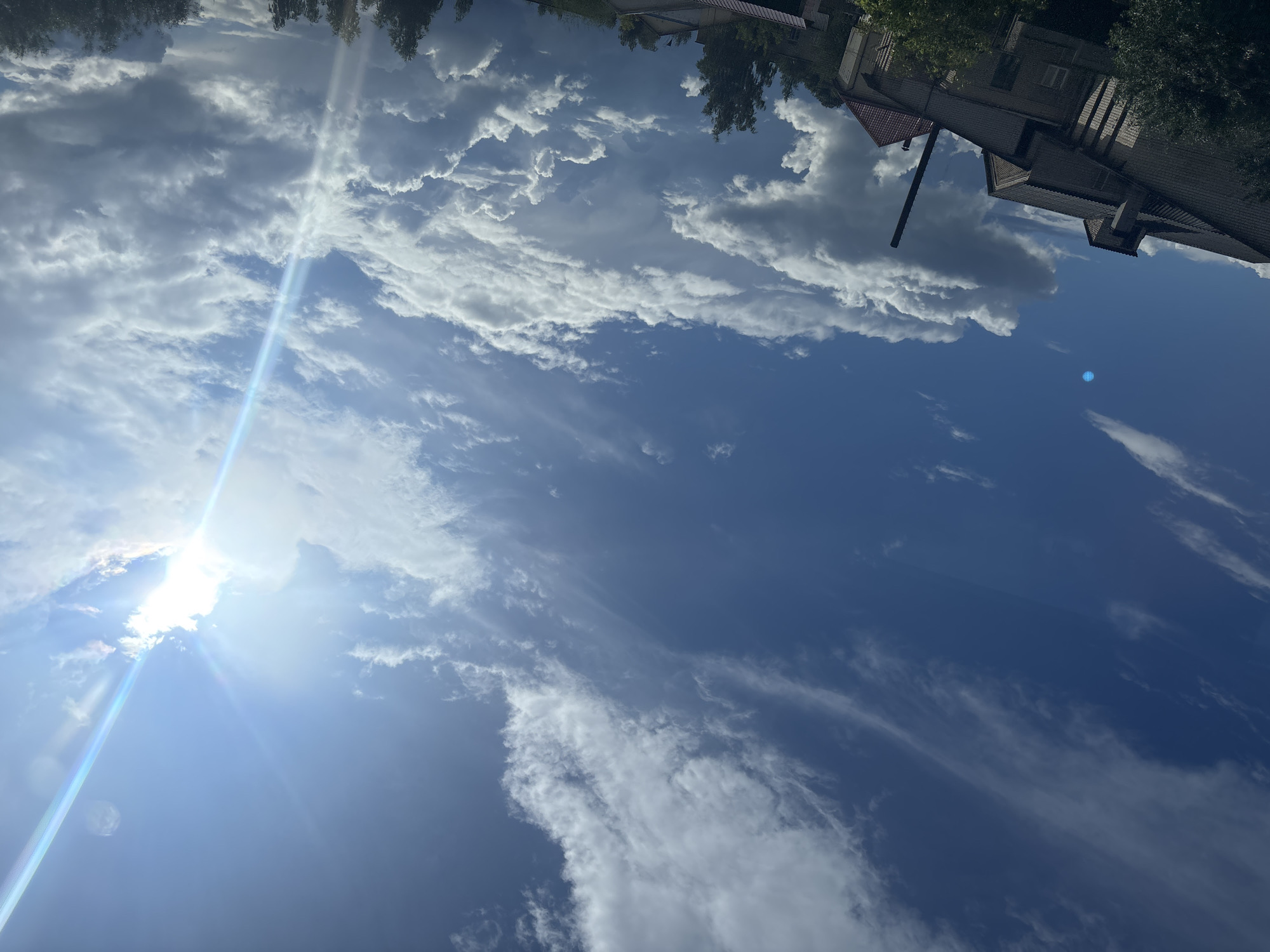 The image depicts a bright, overcast sky with the sun partially obscured by clouds, creating a visible lens flare effect extending diagonally from the upper right corner. The sky is a deep blue with wispy and cumulus clouds scattered throughout. In the lower portion of the image, the rooftops of several buildings and the tops of trees are visible. The architecture appears to be residential or possibly industrial, with brickwork and tiled roofs. The overall impression is that of a sunny day with dynamic cloud formations, taken from a slightly elevated perspective looking upwards. The location is identified as Kapyĺ, Belarus.