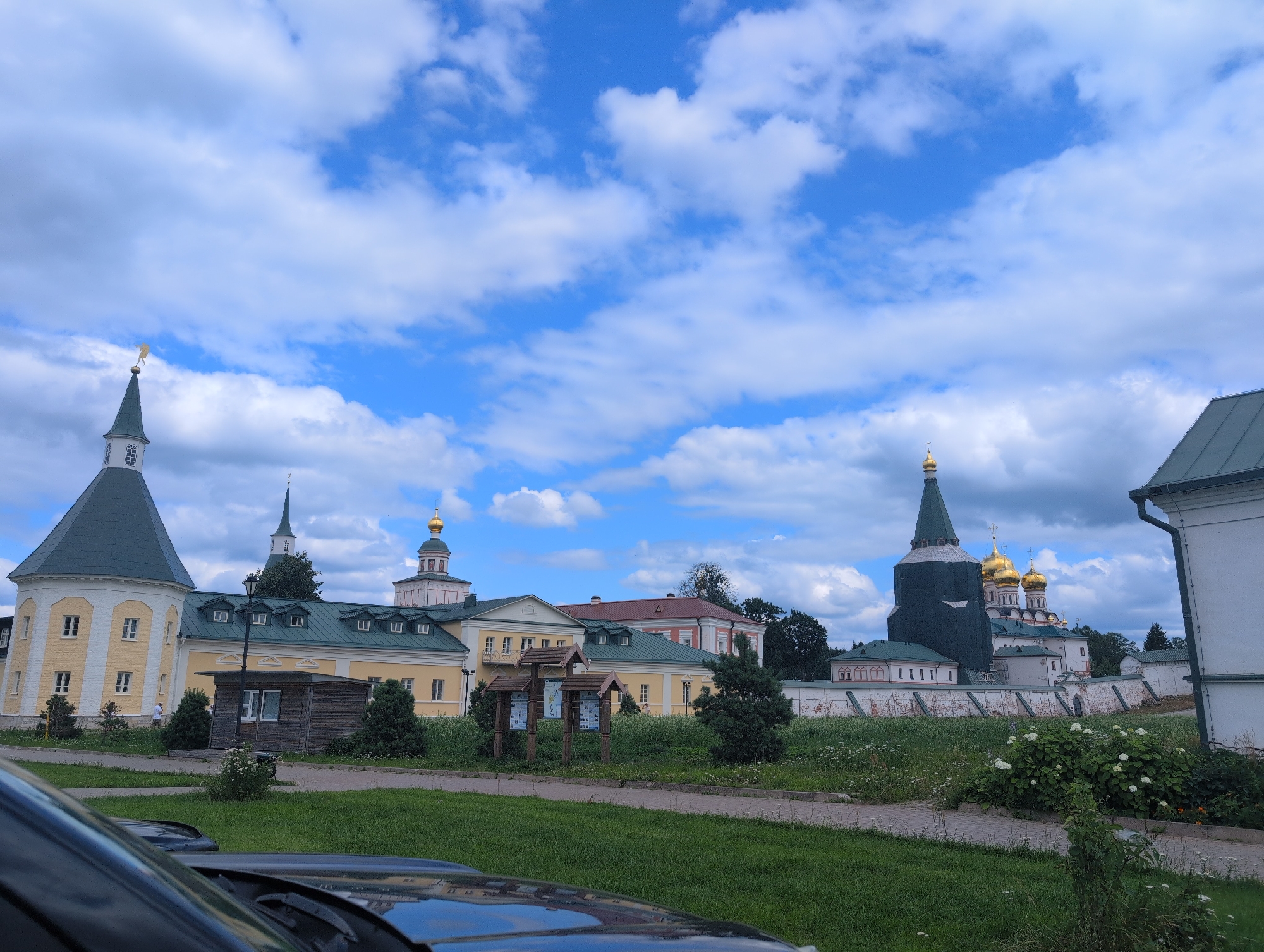 The image captures a serene, sunlit day at a historical religious complex, likely a monastery, under a bright blue sky dotted with fluffy white clouds. The architecture is predominantly Eastern Orthodox, with multiple onion-domed structures, predominantly gold and white, standing against the vibrant sky. The foreground features a lush green lawn with a paved pathway. To the left, a dark car is partially visible, its windshield reflecting the surrounding scene. A small wooden information kiosk stands on the pathway, accompanied by a few trees and shrubs. Further back, a series of buildings with pitched, green roofs and light-colored facades stretch across the frame. A prominent, tall tower with a green conical roof and a golden weather vane stands to the left, while a larger, more elaborate structure with multiple golden domes dominates the right side of the complex. A whitewashed brick wall with arched openings encircles a portion of the complex. In the background, a dense line of trees suggests a natural, possibly wooded, environment surrounding the site. The overall atmosphere is peaceful and picturesque.