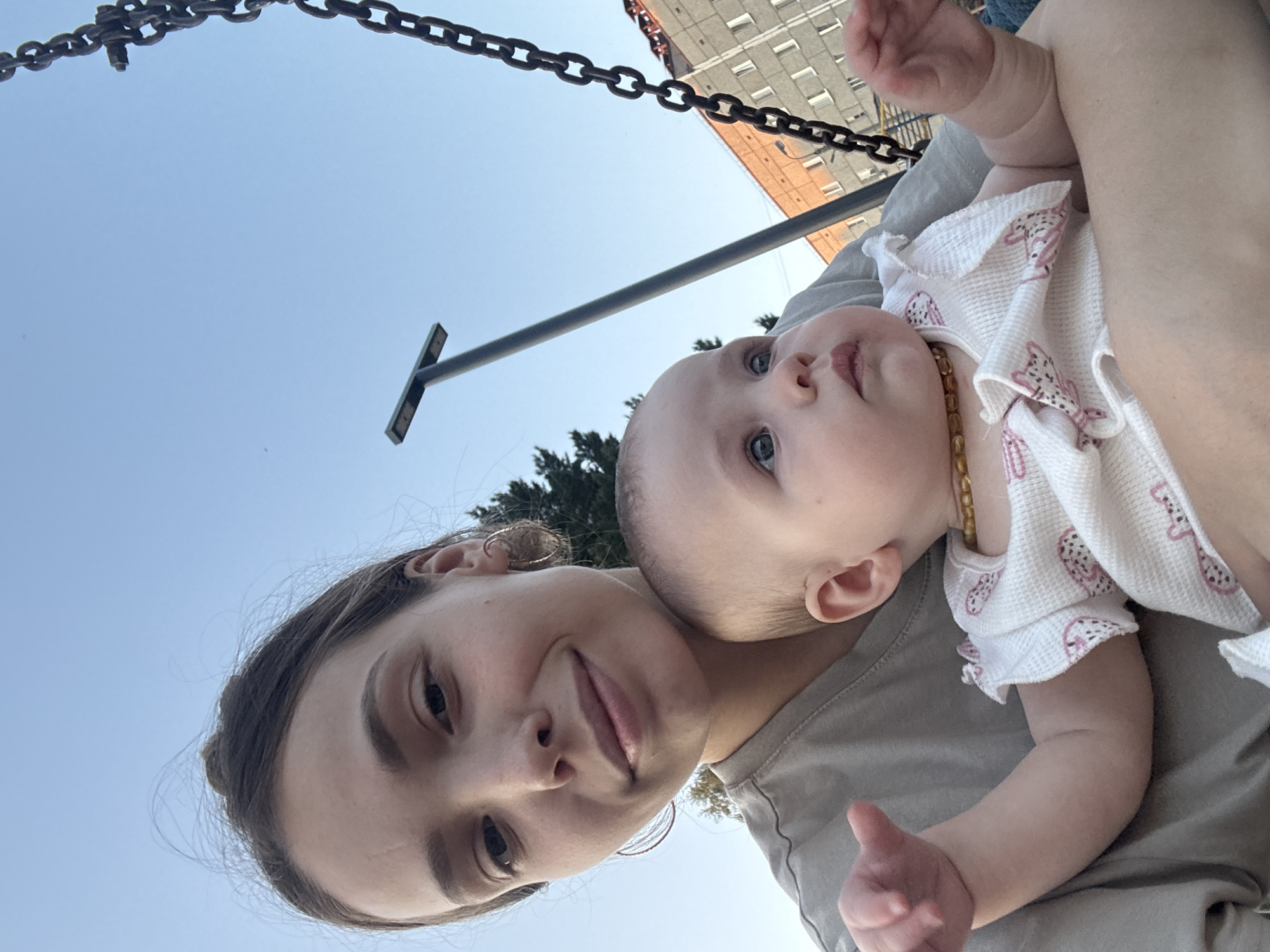 A medium shot captures a young woman holding a baby on a swing. The woman is on the left, with the baby in her arms on the right. The woman has dark hair pulled back and is wearing a grey t-shirt. She is looking at the camera with a slight smile. The baby, facing to the right, has fair skin and light blue eyes, and is wearing a white onesie with a pink pattern. Behind them, the sky is a clear blue. To the right, a large metal chain, part of the swing set, hangs down. In the background, a tall street lamp is visible, and further in the distance, a multi-story brick building stands. The image appears to be taken outdoors in an urban environment, possibly a playground in Noviy, Russia.
