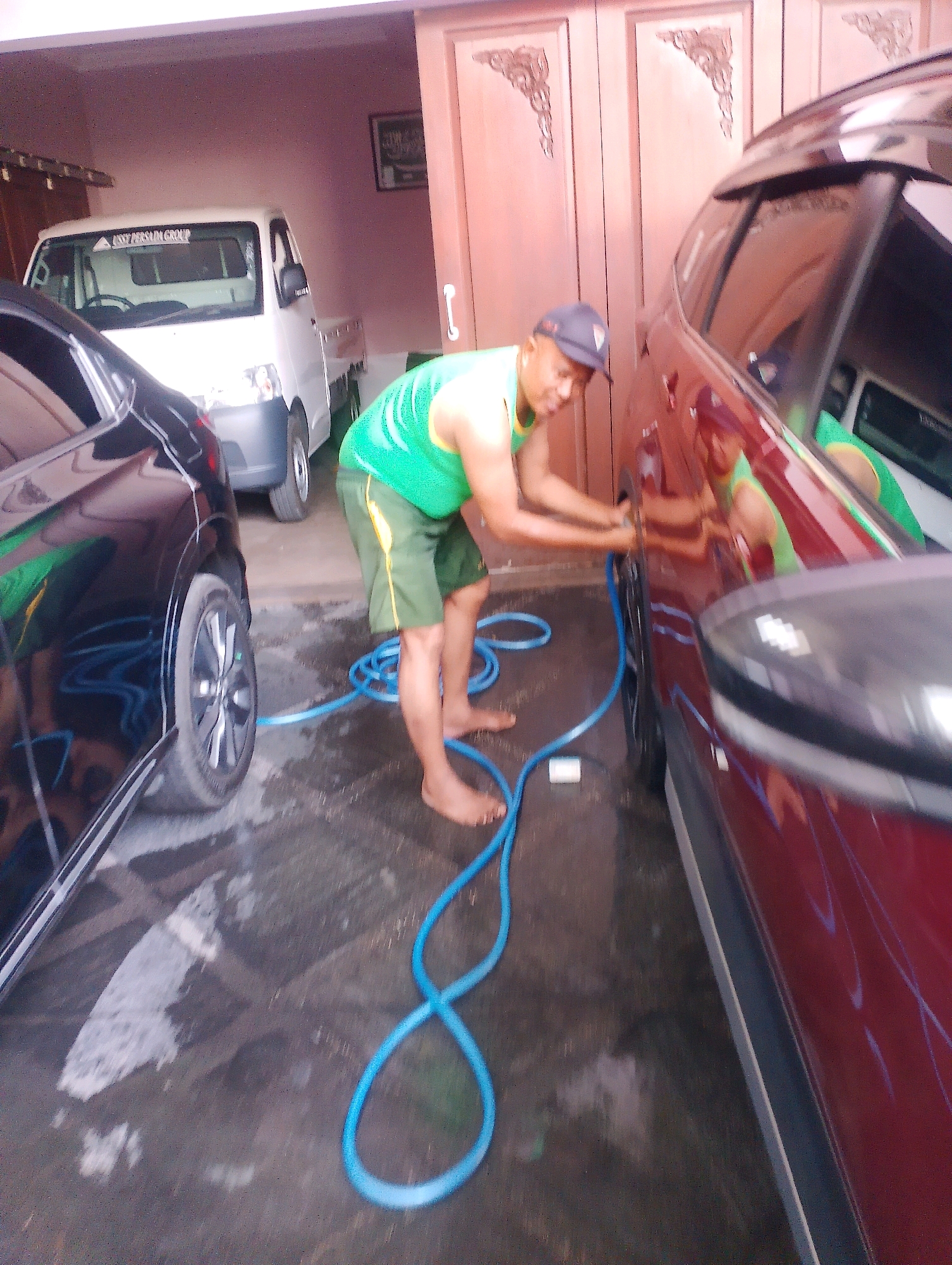 In Probolinggo, Indonesia, a person is washing a maroon car in what appears to be a garage or carport. The individual is wearing a green tank top and green shorts, with a baseball cap on. They are holding a blue hose and directing water towards the car's wheel area. A black car is parked to the left of the maroon car, and a white utility vehicle is visible further back in the garage. The ground is wet, indicating the washing activity. The environment suggests a residential or commercial property with a tiled floor and wooden doors.