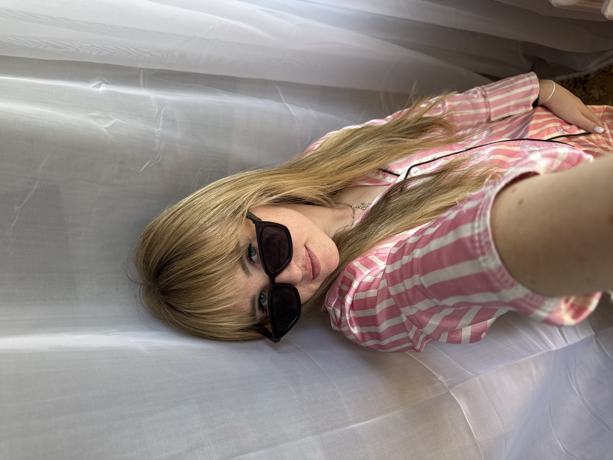 A woman lies on a white cloth wearing sunglasses and a pink and white striped pajama top. Her blonde hair is straight with bangs. She appears to be taking a selfie. The location cannot be determined to be Марковский, Russia from the available visual information.