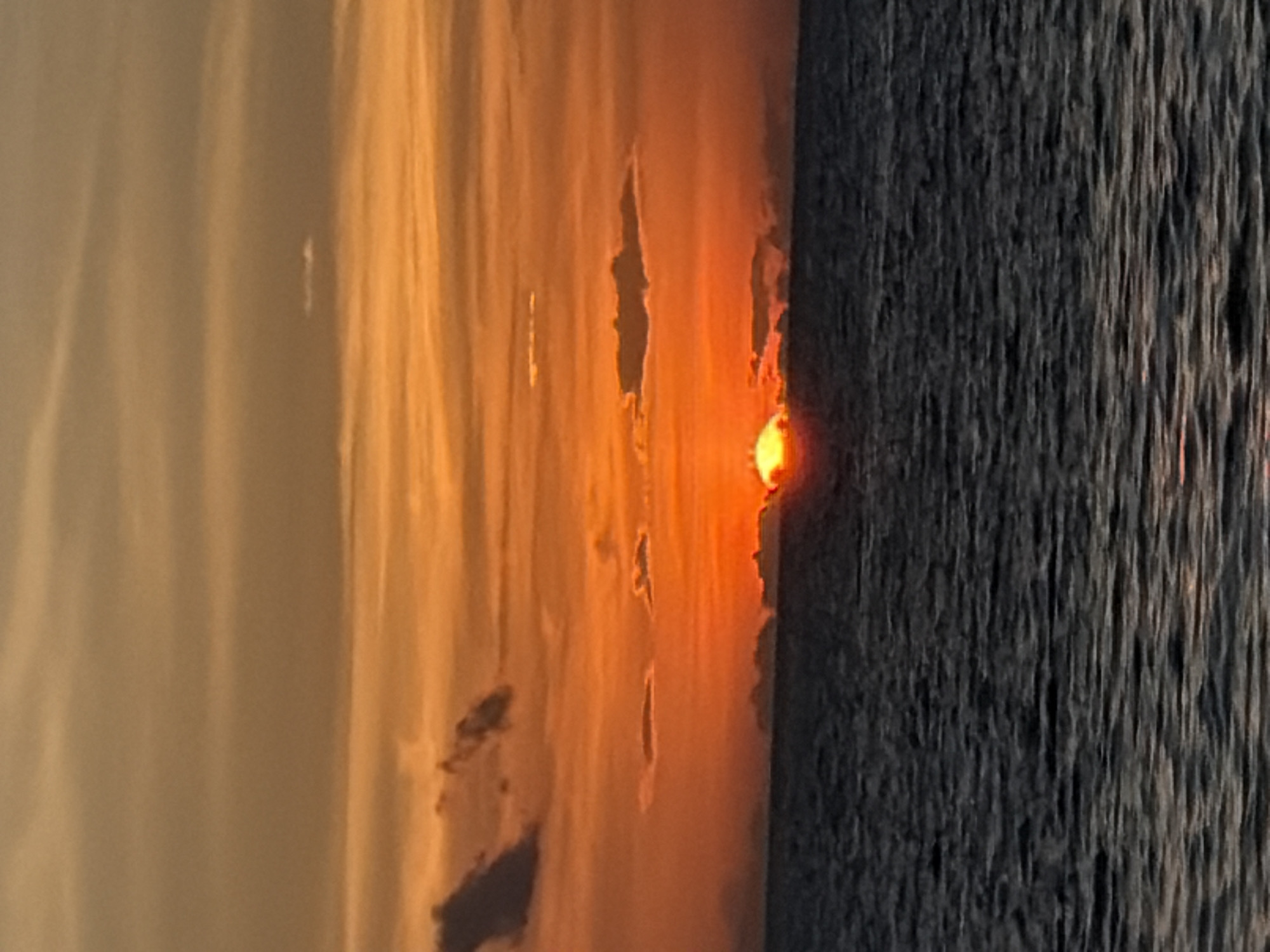 The image depicts a sunset over the ocean in Baan Taling Ngam, Thailand. The sky is filled with vibrant orange and yellow hues, with streaks of darker orange clouds scattered across. The sun is low on the horizon, partially obscured by a thin layer of clouds, casting a warm glow over the scene. The ocean is dark and choppy, reflecting the colors of the sky. The horizon line is clearly visible, separating the sky from the sea. There are no people or man-made objects visible in the image.