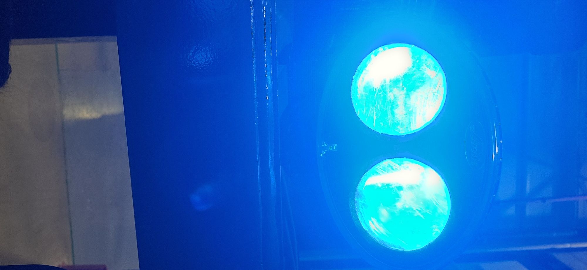 The image captures an indoor scene in Plovdiv, Bulgaria, bathed entirely in a strong, artificial blue light. The dominant features are two vertically stacked, circular light fixtures positioned on the right side of the frame. These lights emit a very bright, almost white-blue glow, indicating powerful illumination.
To the left, a large, lighter-colored rectangular surface, possibly a wall or panel, is visible, reflecting the intense blue light. A subtle vertical green-tinged line runs down this surface. In the bottom-left corner, a dark, indistinct silhouette, possibly the back of a person's head or shoulder, is partially visible.
The blue background surrounding the lights appears to be a solid surface, possibly part of a stage or equipment. Faint textual elements are discernible near the light fixtures; a barely legible, embossed-like word "Arista" can be seen on the casing to the right of the upper circular light in some crops.
No specific activity is taking place, but the powerful, colored lighting suggests a setting like a concert hall, nightclub, or a technical installation where such illumination is used to create a specific mood or for signaling. The time of day is not evident due to the artificial lighting, but the scene is clearly indoors.