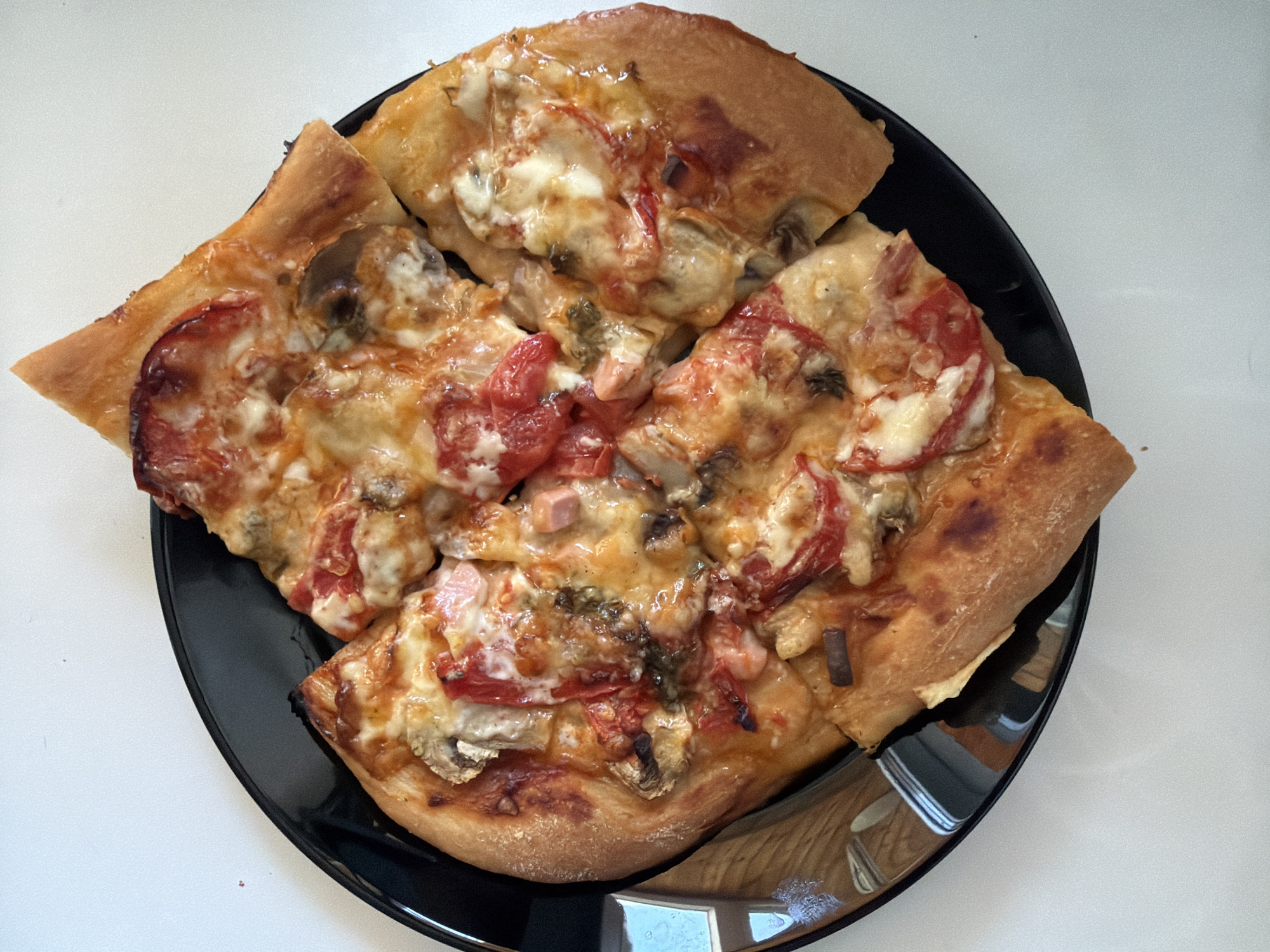 A top-down shot shows four slices of pizza on a black plate. The pizza has a thick crust, and is topped with melted cheese, tomato slices, mushrooms and other ingredients. The plate is on a white surface, possibly a table.