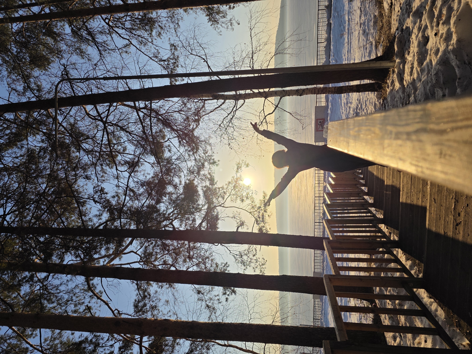 A person stands with arms outstretched on a wooden railing, silhouetted against a bright sun partially obscured by trees. The railing overlooks a landscape of snow and what appears to be a frozen body of water. In the background, a railing runs along the edge of the water. The location is Chaikovsky, Russia, evidenced by the image's origin. The scene is framed by trees, some with bare branches, against a blue sky. A wooden staircase leads down to the snow-covered ground.