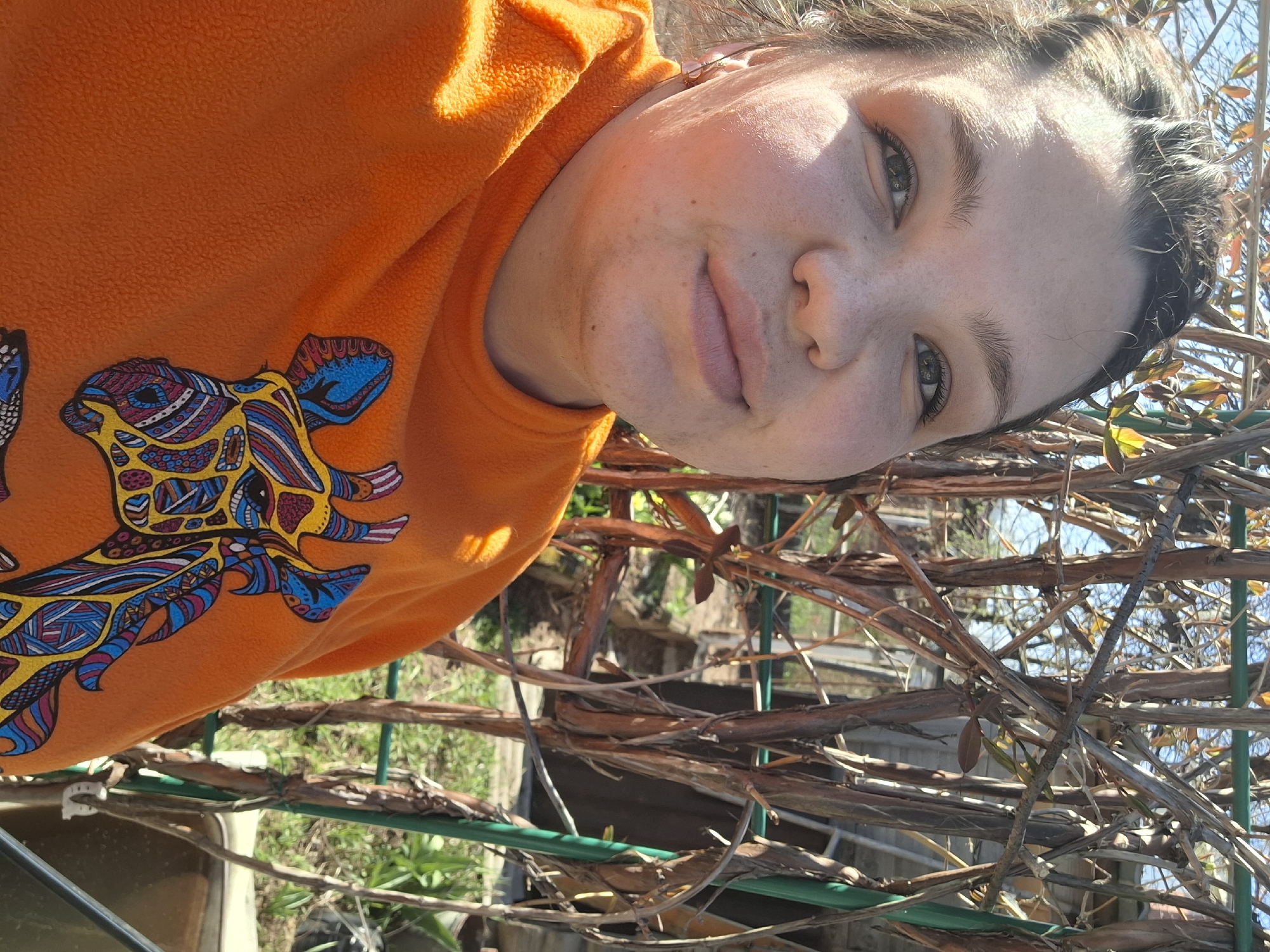 A young woman is pictured from a high angle in what appears to be an outdoor setting. She is wearing an orange fleece sweatshirt adorned with a colorful abstract design. Her face is visible and partially illuminated by sunlight. The background includes a structure made of interconnected branches and green supports. The environment appears to be a garden. The location is stated to be Chaikovsky, Russia, but this is not explicitly verifiable from the image content.