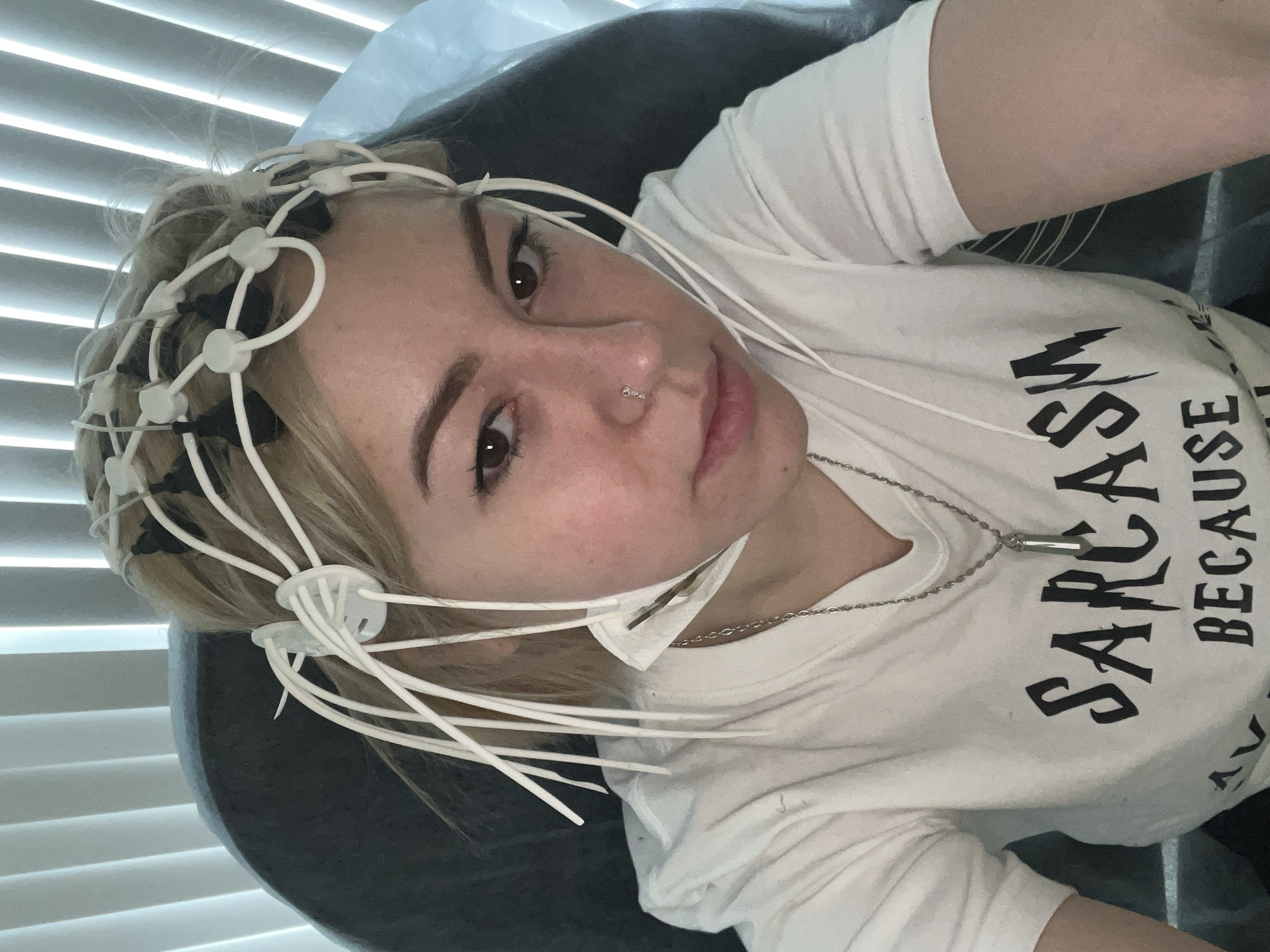 The image captures a young person, likely in Odesa, Ukraine, reclining and wearing an electroencephalography (EEG) cap. This medical device, a white flexible framework with multiple electrodes, is fitted onto their blonde hair and scalp, with wires extending down. The subject looks directly at the camera with a neutral expression.
Notable features include their dark eyes, defined eyebrows, and a small hoop piercing in their left nostril. They are wearing a white short-sleeved t-shirt with black text on the front, partially visible as "SARCASM BECAUSE". A silver-colored chain necklace with a cylindrical pendant hangs around their neck.
The setting appears to be indoors, likely a medical facility or research lab, given the specialized equipment. Horizontal blinds are visible in the background, suggesting a window and controlled lighting. The person is resting against a dark headrest or chair. The activity taking place is the recording of brain activity, possibly for diagnostic or research purposes.