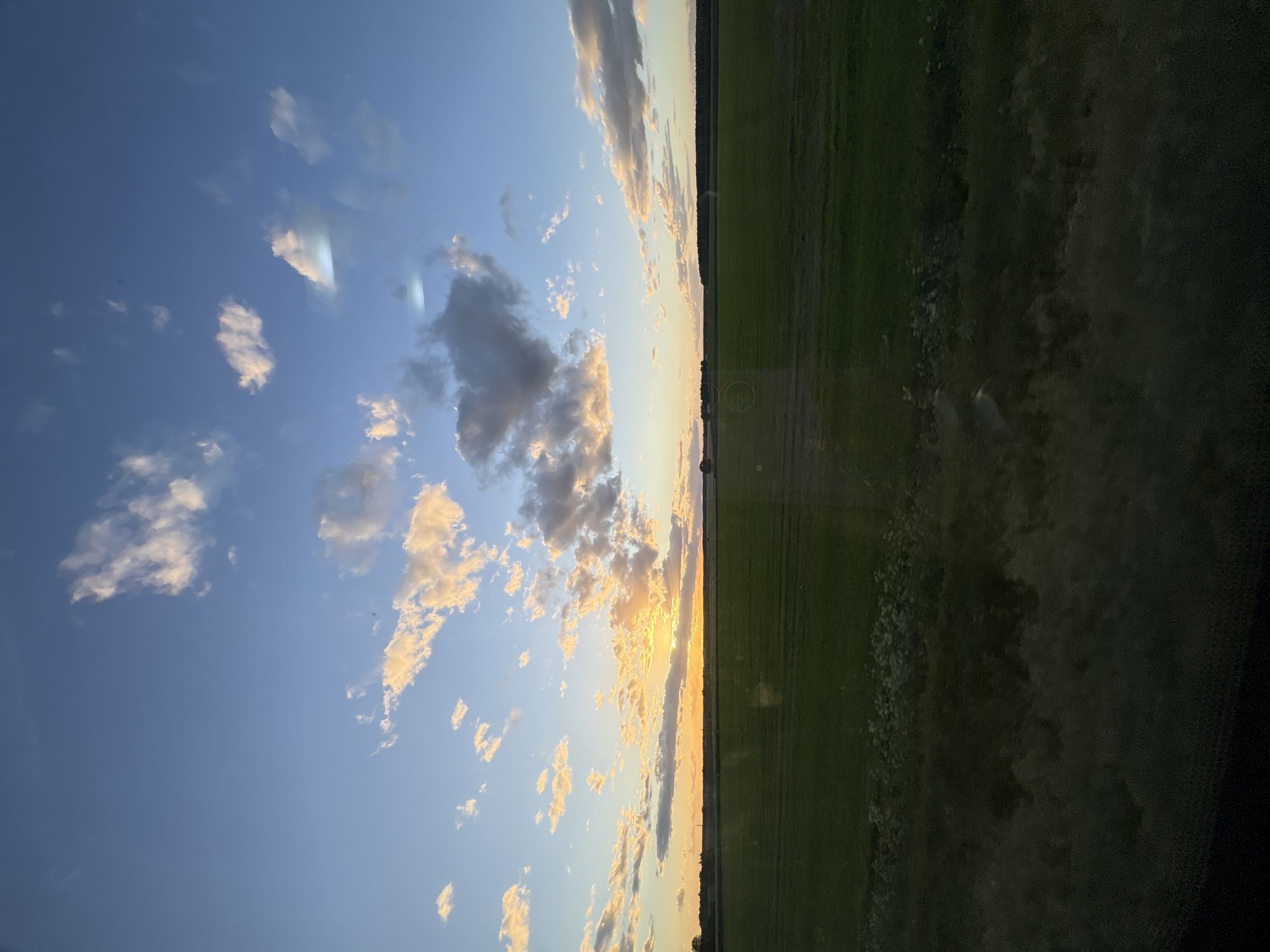 The image depicts a landscape in Мажэйкаўскі сельскі Савет, Belarus, split between a sky and a field. The sky, primarily blue, is populated with scattered, sunlit clouds, particularly dense towards the horizon. The field appears green and stretches towards the horizon, bordered by a line of trees. The sunset casts a golden hue on the clouds and the horizon.
