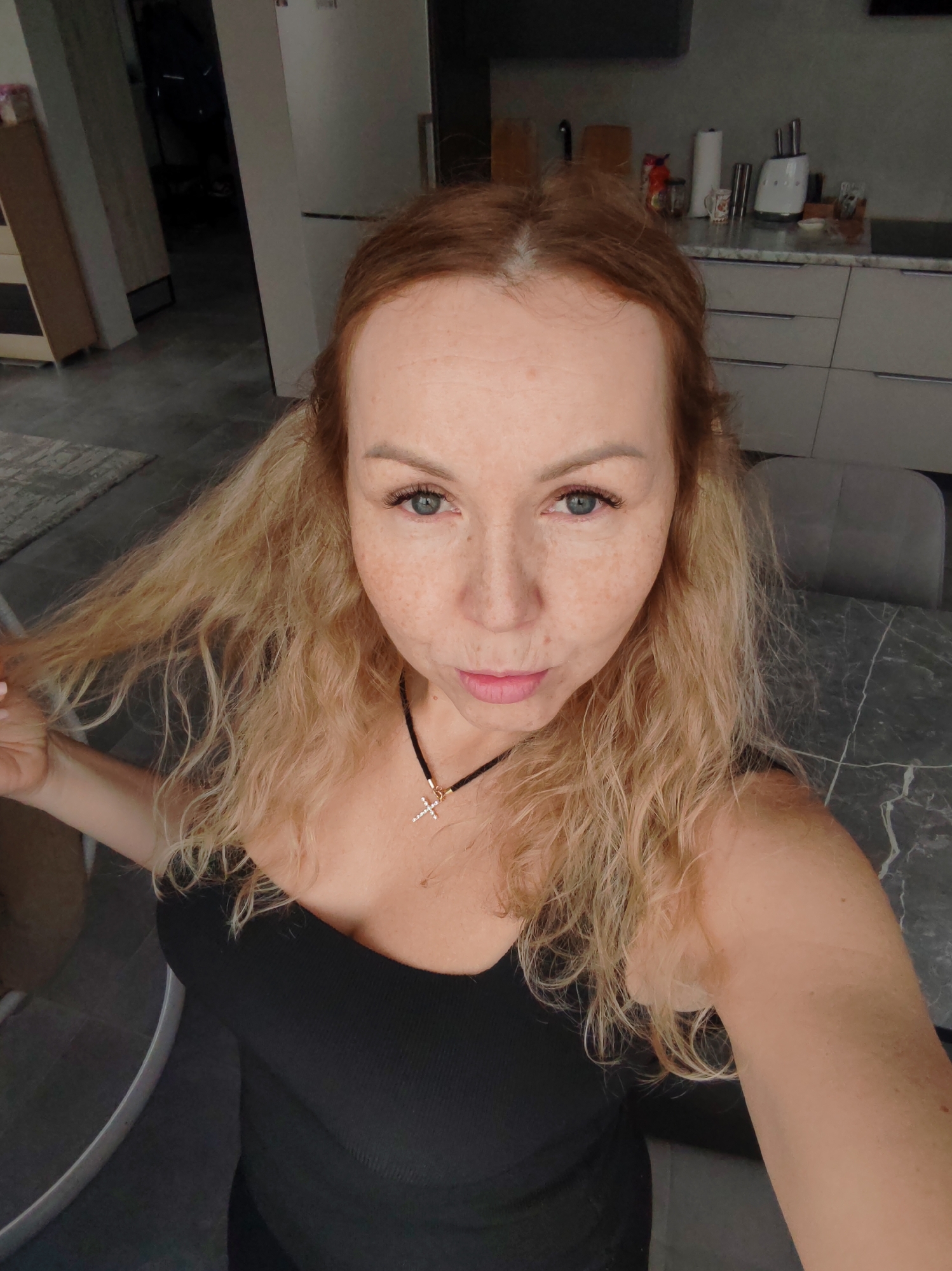 A fair-skinned woman takes a selfie indoors in what appears to be a modern home in Chaikovsky, Russia. She has light brown hair at the roots that transitions to wavy blonde ends, and notable freckles across her face. Her blue eyes look directly at the camera, and her lips are slightly pursed. She is wearing a black strapless or spaghetti-strap top and a black cord necklace with a small, sparkling gold-colored cross pendant. With her left hand, she gently holds a strand of her wavy hair.
The background shows a contemporary interior. To her right, a dark grey marble-patterned table is visible, alongside the back of a grey chair. Behind her, a modern kitchen area features light-colored cabinets with silver handles and a light countertop adorned with a paper towel roll, a mug, and small kitchen appliances. Further back and to her left, a grey floor with a lighter grey rug and part of a white door or cabinet are visible, leading into a dimly lit hallway or room. The overall lighting suggests it is daytime. No text is visible in the image.