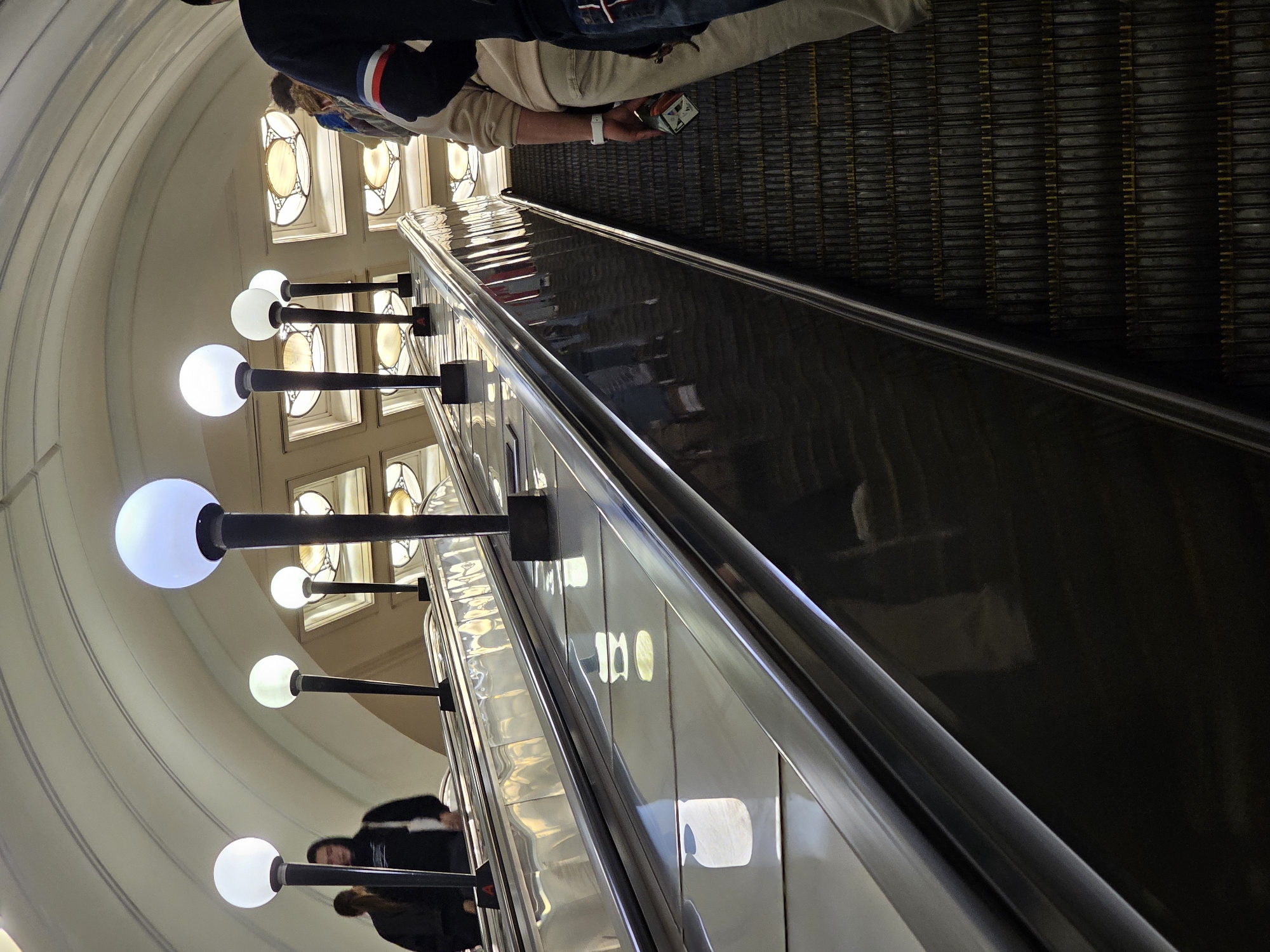 The image depicts an interior scene inside a Moscow Metro station. Two escalators are visible, angled upwards towards the top of the frame. The escalators have metal treads and handrails. Along the side of the escalator on the left, there are several spherical lights mounted on dark poles. These lights are illuminated, casting a soft glow. Above the escalators, a vaulted ceiling is visible, with decorative panels and recessed lighting. On the escalators, several people are ascending. On the left escalator, two individuals are visible, one wearing a dark hoodie and the other in a dark jacket. On the right escalator, a person in a light-colored sweatshirt and beige pants is holding a small object in their hand. The overall atmosphere is clean and well-lit, characteristic of a metro station.