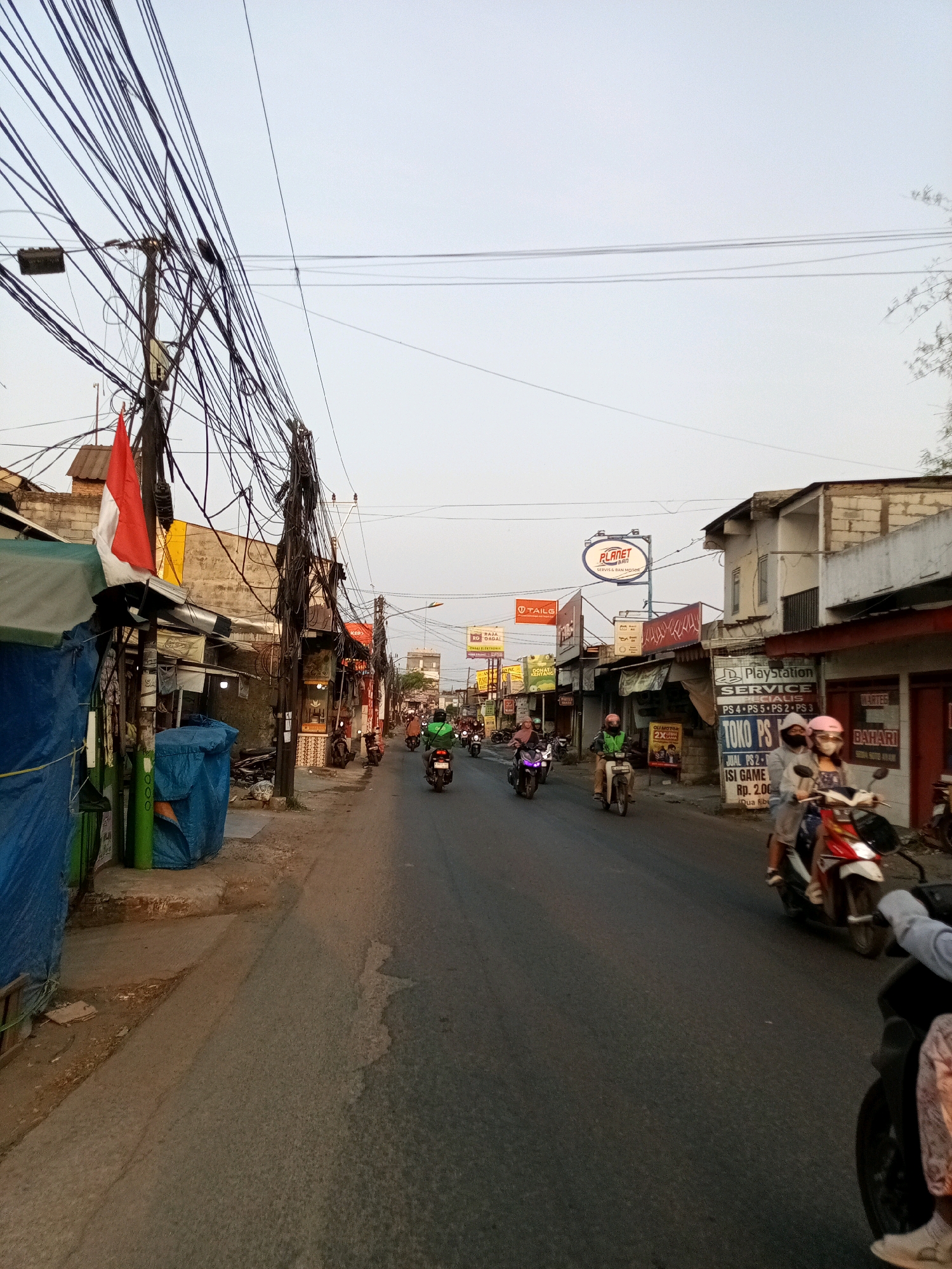 The image depicts a street scene in Karangsatria, Indonesia, characterized by a dense overhead network of electrical wires. The street is paved and appears to be a busy thoroughfare with several motorcycles navigating the road. On the left side of the street, there are small shops and market stalls, some covered with blue tarpaulins. An Indonesian flag is visible, mounted on a pole near one of the stalls. On the right side of the street, there are more shops, including one with a "Playstation" sign and another advertising "BAHARI". The sky is overcast, suggesting it is either early morning or late afternoon. Several individuals are visible riding motorcycles, some wearing helmets. The overall impression is one of a bustling, urban environment with a focus on commercial activity and daily life.