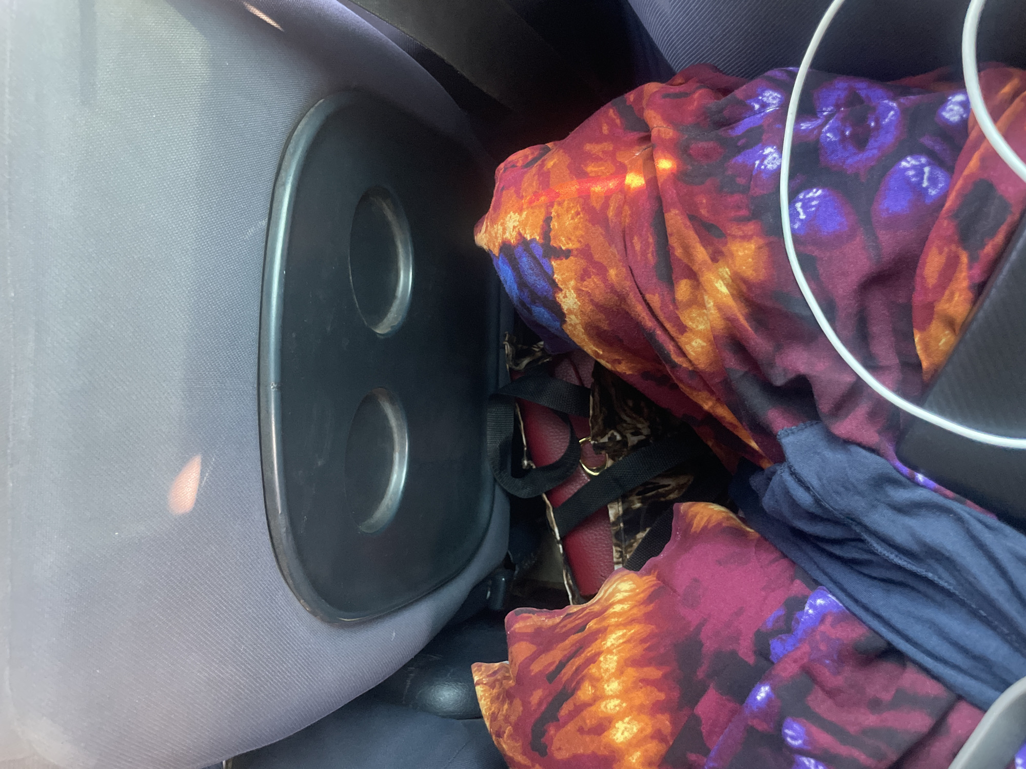 A high-angle close-up shot shows the interior of a vehicle, likely a car, in Wauro Jabbe, Nigeria. The focus is on the back of a seat, which is upholstered in grey fabric and features a black tray with two circular indentations. Underneath the tray, various items are piled: a red handbag with black straps and gold hardware is partially visible, along with a leopard-print bag. In the foreground, two brightly colored garments are folded and stacked. One is a predominantly red and orange patterned fabric with swirls of purple, and the other is a dark blue folded garment. A white circular object, possibly part of a car accessory or personal item, is visible at the bottom right. The lighting suggests it is daytime, with some sunlight reflecting off the objects. No people are visible in the image.