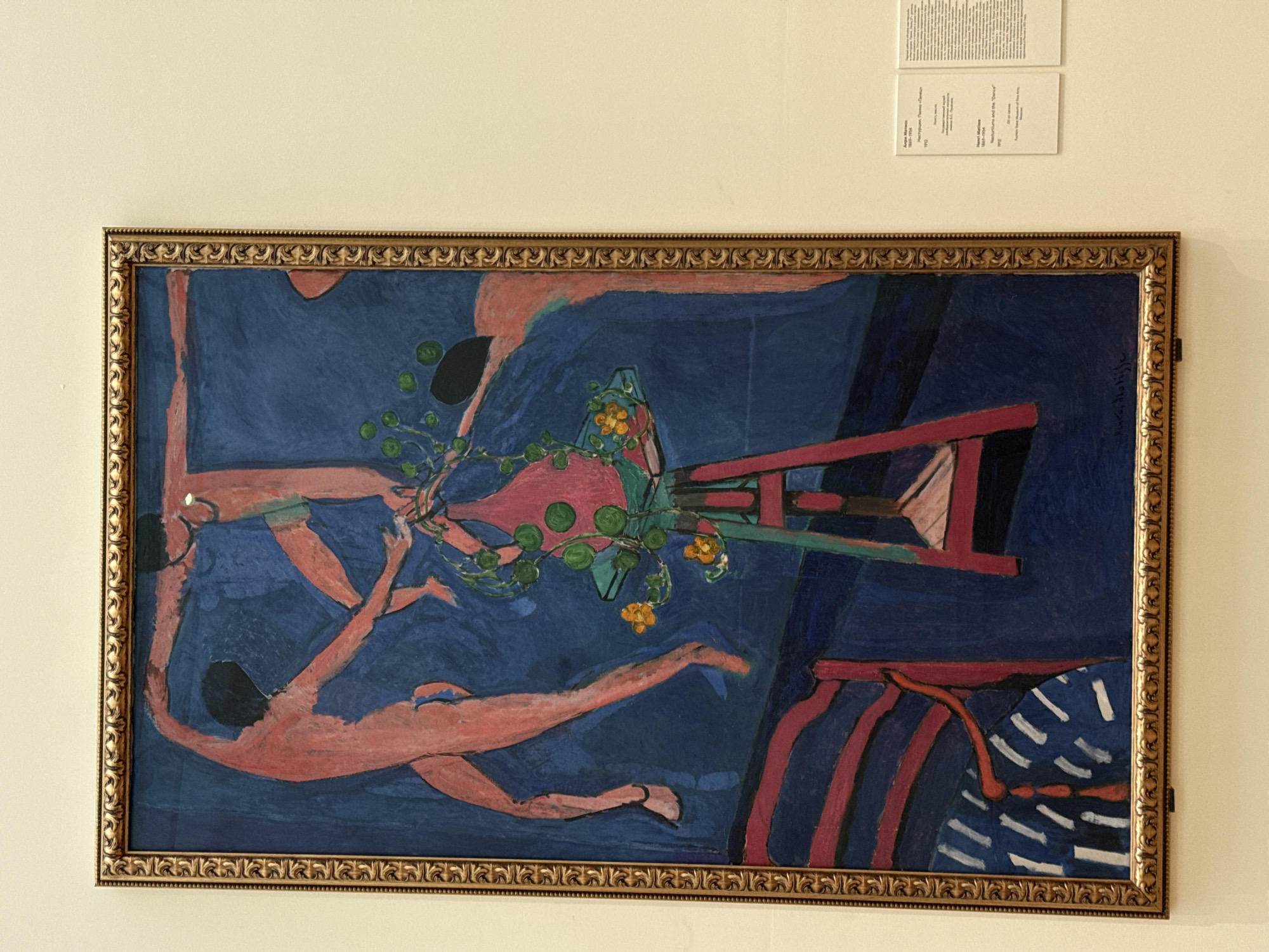 A framed painting hangs on a neutral-toned wall. Two printed cards are placed above the painting. The painting itself features figures with pink-toned skin in a blue-dominated scene. The painting's subject is "Naturmort and the 'Dance'," an oil on canvas by Henri Matisse, 1912, from the Pushkin State Museum of Fine Arts in Moscow, Russia. The figures appear nude and are positioned around a vase or still life arrangement. There are shapes of black, green, and yellow with red and white stripes present. The painting is in an ornate, gold-colored frame.