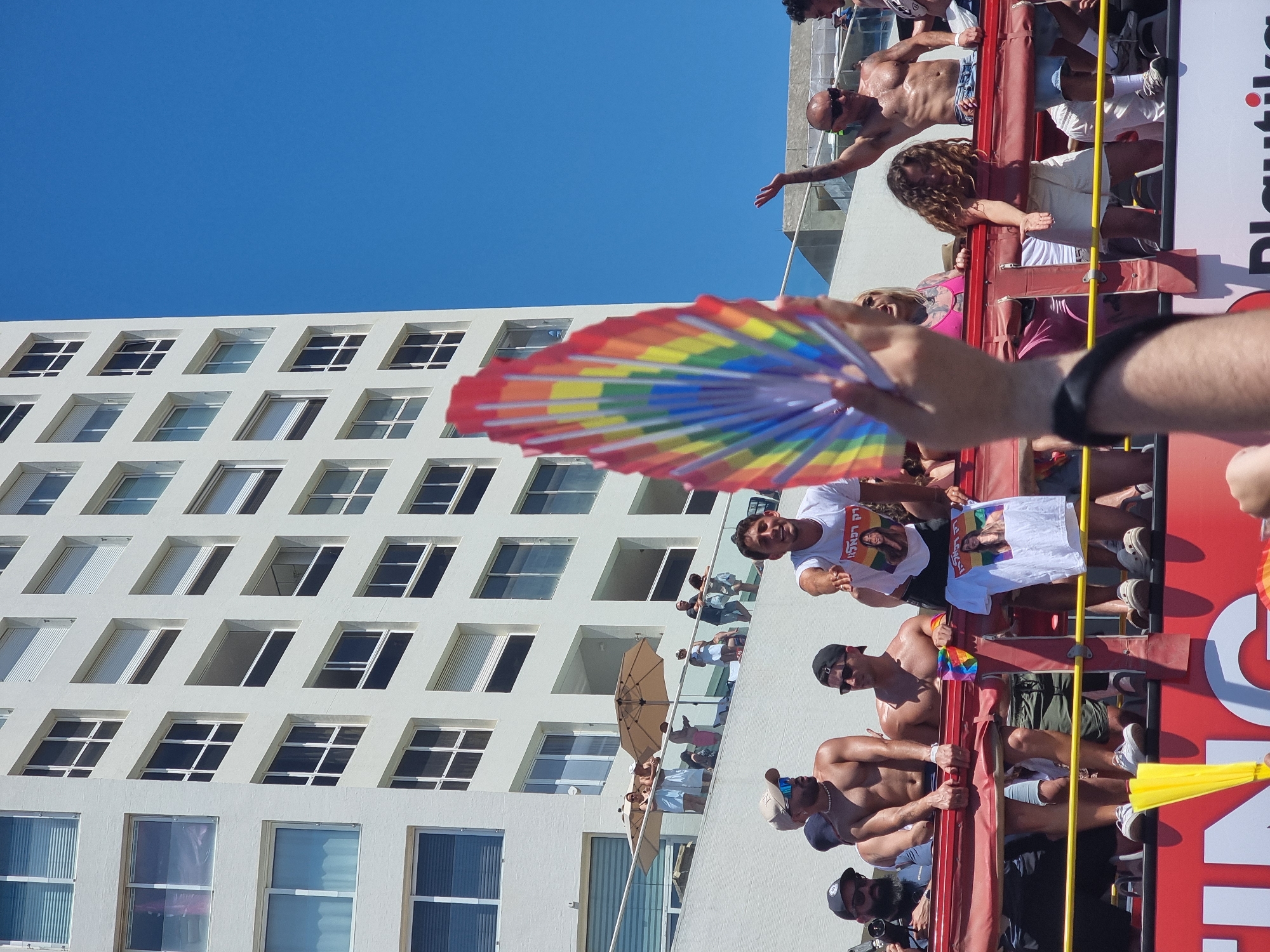 This is a photograph taken at a Pride parade in Tel Aviv, Israel. The scene is dominated by a tall, white, modern building with many windows, and a bright blue sky. In the foreground, people are visible on what appears to be a float or elevated platform. One person is holding up a rainbow-colored fan. Another person is wearing a white t-shirt with a picture of a woman on it, and text that appears to be in Hebrew. Several shirtless men are standing and sitting on the platform. Other people are visible in the background, some on balconies of the building and others on the street level. The atmosphere suggests a festive and celebratory event.