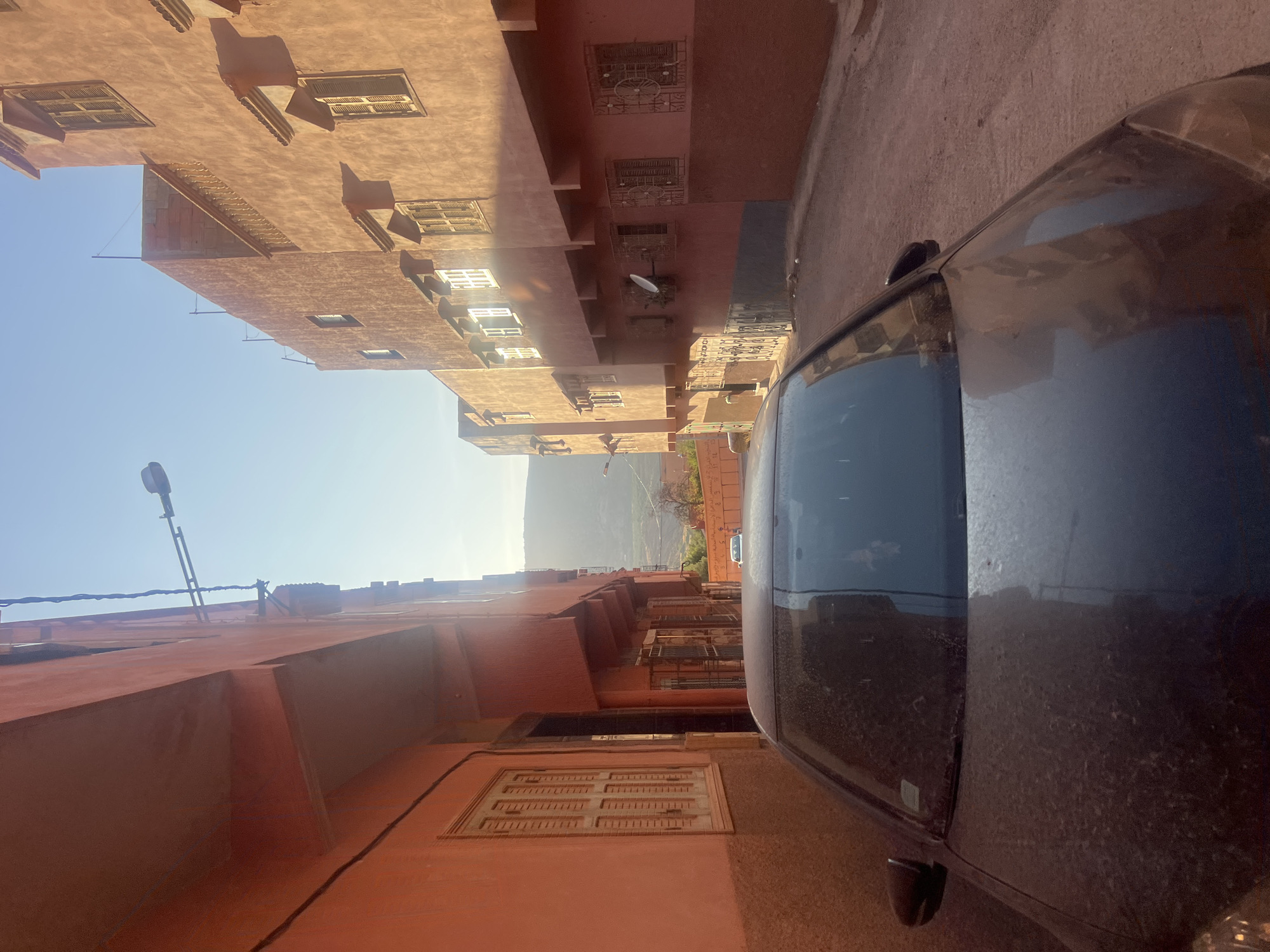 The image depicts a narrow street in Ouaouizert, Morocco, with characteristic terracotta-colored buildings lining both sides. The architecture suggests residential structures with multiple stories, featuring wooden shutters on the windows and decorative architectural elements. The street itself is unpaved, with a dusty, sandy surface. The lighting indicates either early morning or late afternoon, with long shadows cast by the buildings and the sun low in the sky, creating a warm, golden hue over the scene. A dark-colored car is parked in the foreground, partially obscuring the view of the street ahead. In the distance, the street opens up to reveal a hilly landscape under a clear blue sky. The overall impression is one of a quiet, somewhat dusty urban environment typical of smaller towns in the region.