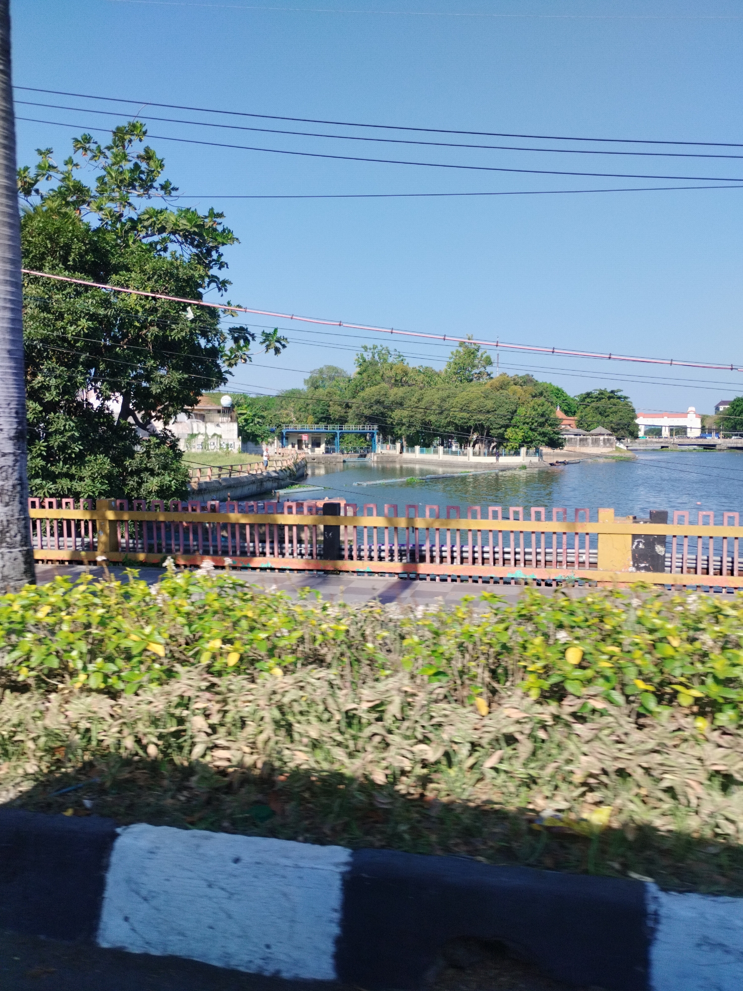 The image depicts a sunny day scene of a park with a body of water, likely a lake or reservoir, in Surabaya, Indonesia. A yellow and red striped fence runs along the edge of the water, separating it from a grassy area and a road. In the background, there are trees, buildings, and some infrastructure related to water management. The sky is clear and blue, with several power lines crisscrossing the upper part of the frame. The overall impression is one of a public recreational space.
