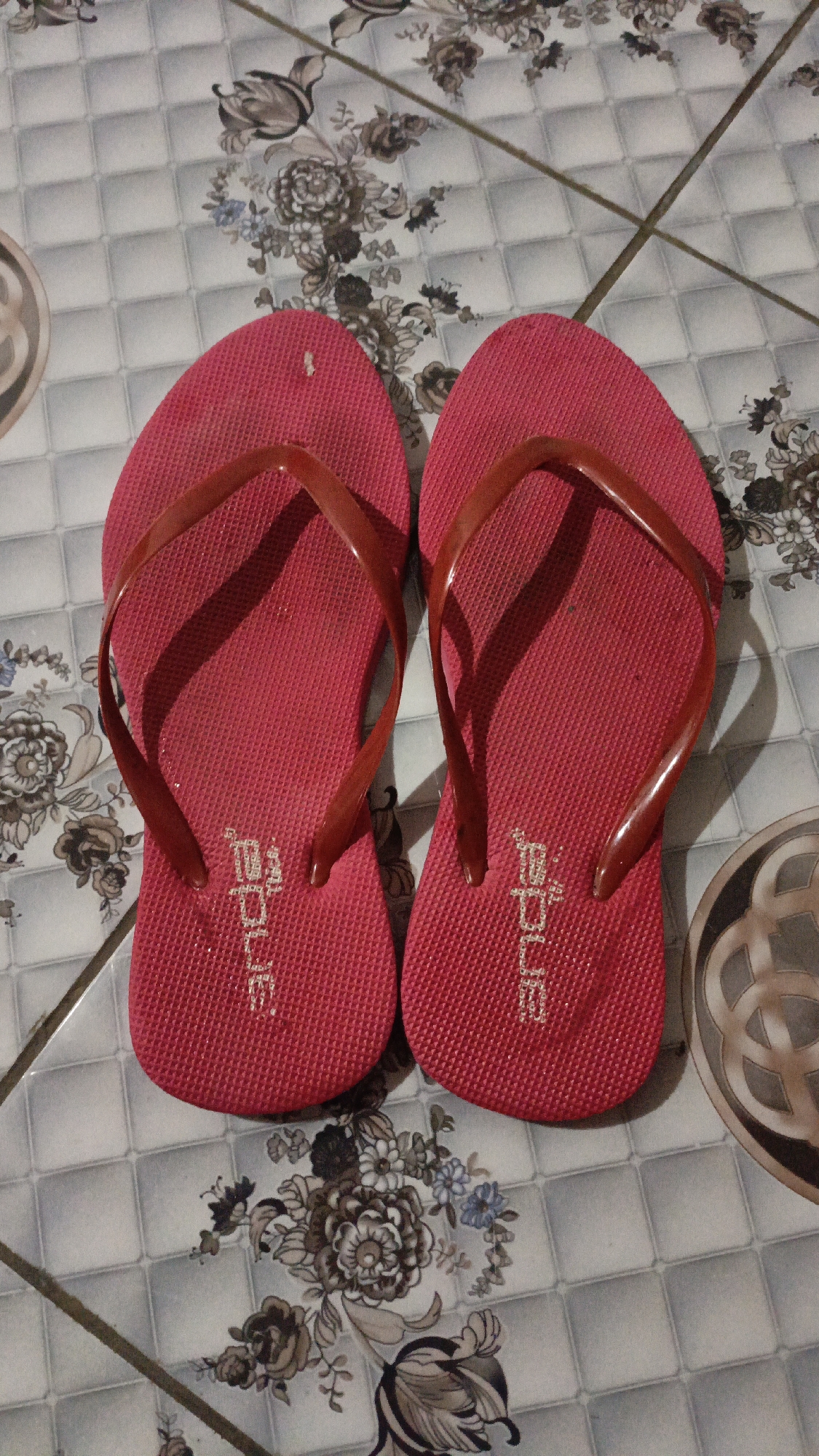 The image displays a pair of red flip-flops lying on a tiled floor. The tiles have a light grey grid pattern with floral motifs in shades of grey and brown. The flip-flops are positioned side-by-side, with the straps facing upwards. The brand name "Bata" is visible in white lettering on the sole of each flip-flop. The lighting suggests the image was taken indoors, possibly in a home or a public restroom, given the tile pattern. There is no indication of the specific location within Pejala, Indonesia.