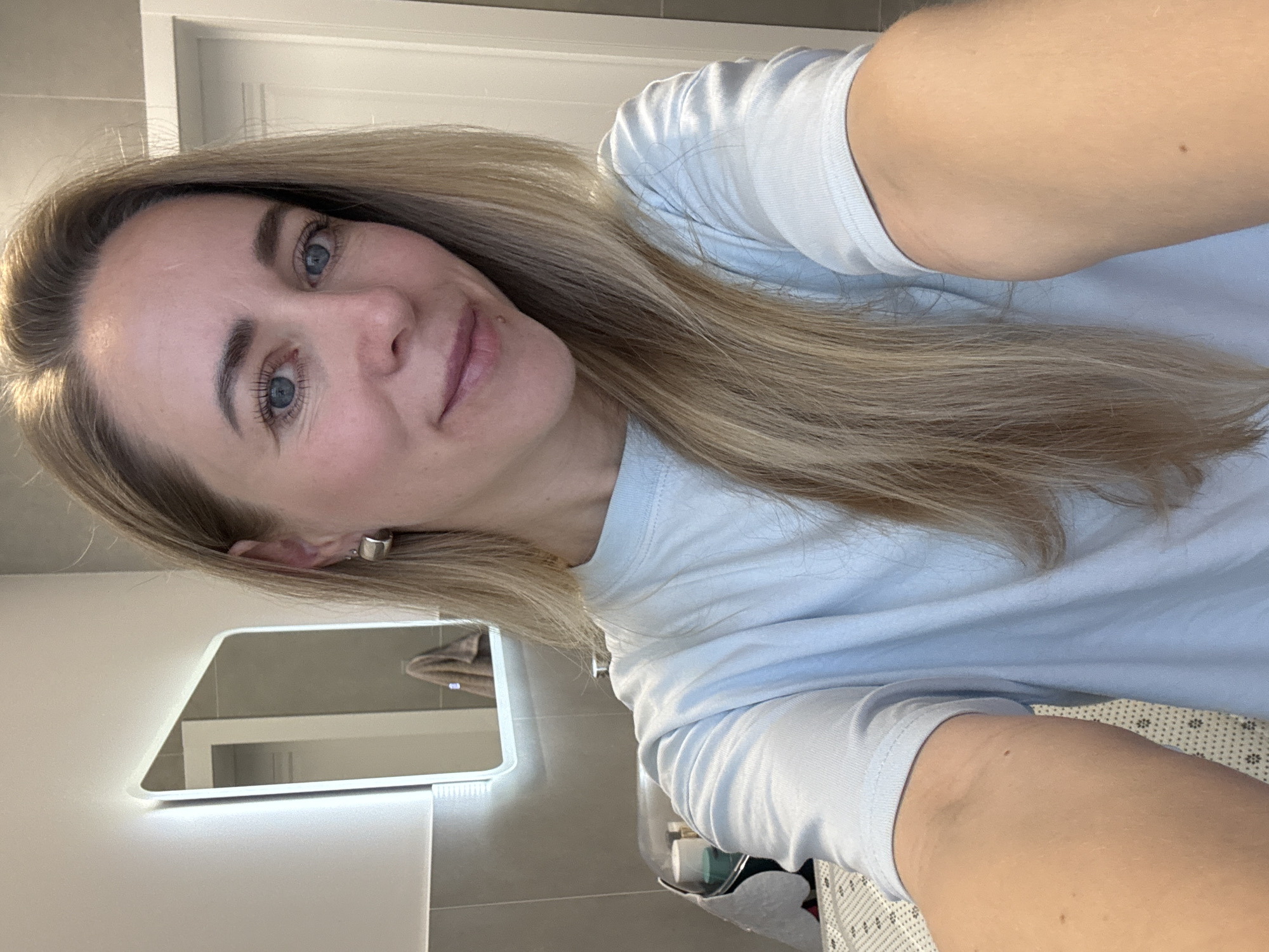 A fair-skinned woman with long, straight blonde hair and blue eyes is pictured taking a selfie indoors in Chaikovsky, Russia. She is wearing a light blue crew-neck t-shirt and has a subtle, pleasant smile directed towards the camera. A small mole is visible above her upper lip, and another on her right upper arm. Her left ear features a small, simple silver stud earring.
The setting appears to be a modern bathroom, with a sleek, rectangular mirror positioned behind her to the left. This mirror features integrated backlighting along its edges, illuminating the space. Reflected in the mirror are light gray or beige tiled walls and a white doorframe. The overall lighting is bright and even, suggesting indoor artificial light. The reflection in the lower right portion of the mirror hints at other bathroom fixtures or items on a counter. No visible text is present in the image.