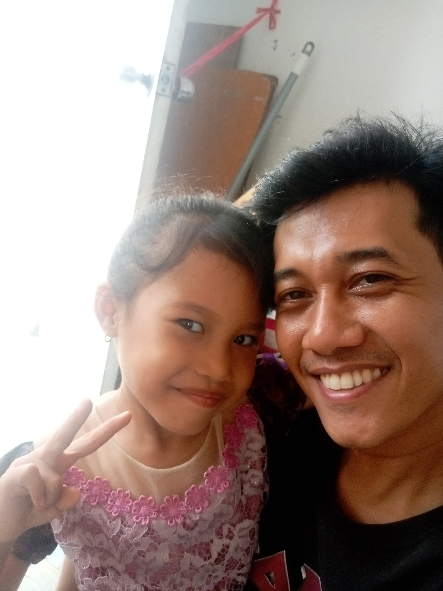 The image is a selfie of an adult male and a young girl, taken from a close-up perspective, likely indoors. The male is on the right, smiling broadly, with short, dark hair and dark eyes. He is wearing a black t-shirt. The girl is on the left, also smiling, with her hair pulled back. She is wearing a light-colored dress with pink floral lace trim around the neckline. Her left hand is raised, making a peace sign. The background is out of focus but appears to be an interior space with a white wall, a door frame, and possibly furniture. The lighting is bright, suggesting it might be daytime. There is no identifiable information in the image that would indicate a specific location within the city of Batam, Indonesia.