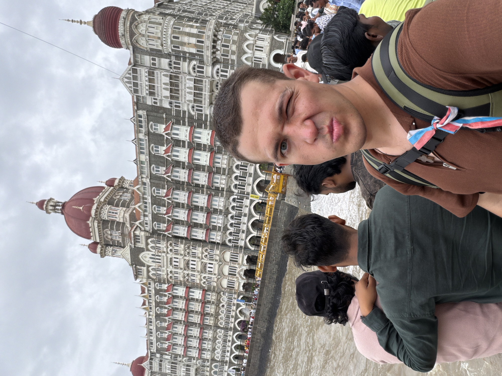 A man winks at the camera in the foreground of this outdoor, eye-level, medium-shot photograph. The man is wearing a brown shirt and a green and black backpack with a red, white, and blue ribbon tied to the strap. Behind him, the Taj Mahal Palace Hotel, a historic luxury hotel in Mumbai, India, is visible. The hotel is a grand, ornate building with red domes and many windows. A body of water, likely the Arabian Sea, is in front of the hotel. Several other people are visible in the background, some standing near the hotel entrance and others in a crowd to the right of the frame. The sky is overcast.