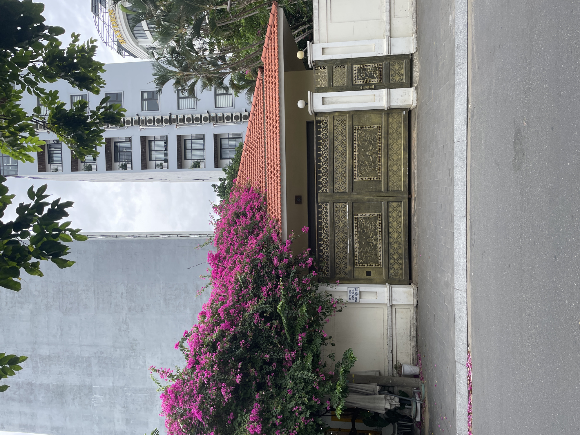 The image depicts a street scene in Đà Nẵng, Vietnam. In the foreground, a paved road is visible, with a gray concrete sidewalk bordering it. Beyond the sidewalk, a house with a red tile roof features an ornate, gold-colored gate. A vibrant bougainvillea bush, heavy with pink flowers, cascades over the roof of the house and drapes down its side. Behind the house, a tall, gray building with a grid-like pattern of windows stands prominently. To the right of the gray building, a lighter-colored, modern building with multiple balconies and air conditioning units is visible. Further to the right, a curved white building with the word "HAYDEN" is partially obscured by palm trees. The sky is overcast. A small sign with Vietnamese text is affixed to the wall next to the gate.