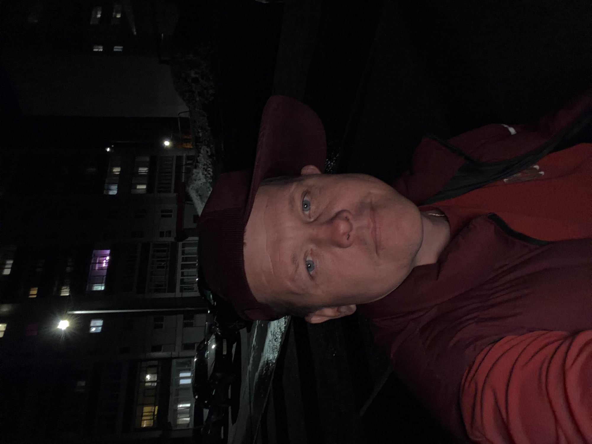 The media file displays a self-portrait, likely a selfie, of a man captured at night. The image is presented rotated 90 degrees clockwise.
**Subjects:** The central subject is a man with blue eyes and fair skin, wearing a dark reddish-brown cap and a red jacket. He has a neutral to slightly serious expression and is looking directly at the camera. In the background, several tall, dark buildings with numerous illuminated windows are visible, suggesting an urban or suburban setting. What appears to be snow or ice covers a railing or dark surface in the mid-ground, located above the man's head in the rotated image.
**Activity or event taking place:** The man is posing for a close-up photograph of himself, seemingly taken outdoors in the evening.
**Notable details:** The time of day is clearly night, indicated by the dark sky and the artificial lights glowing from the building windows. The presence of snow or ice on a nearby surface suggests cold weather, possibly a winter night. The buildings appear to be multi-story residential structures. No visible text is present in the image.