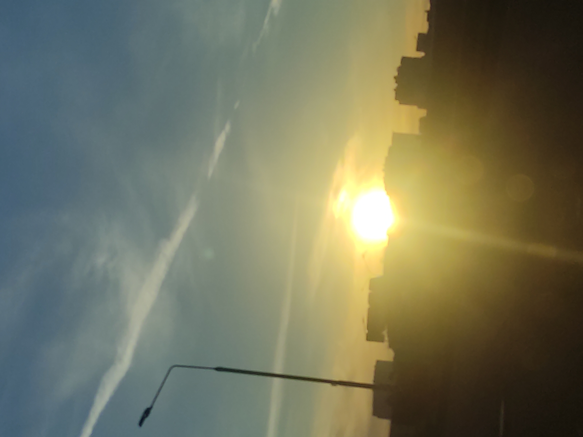 The image captures an urban or suburban landscape bathed in the warm, dramatic light of either sunrise or sunset. The sky dominates the upper portion, transitioning from a deep golden yellow near the horizon where the sun is located, to a lighter blue in the upper left. The sun itself is a brilliant, intense orb positioned on the right side, emitting strong rays and lens flare, creating a significant glare.
Silhouetted against the bright sky are numerous buildings, forming an irregular, dark skyline across the lower half of the image. Their details are obscured by the backlight, rendering them as dark masses. A prominent, dark street lamp pole extends upwards from the lower left, reaching towards the middle of the frame. In the blue part of the sky, subtle, wispy white clouds or contrails are visible, adding texture.
No people or discernible activities are taking place, focusing solely on the atmospheric qualities of the scene. The overall impression is one of peaceful urban beauty at the start or end of the day. There is no visible text in the image.