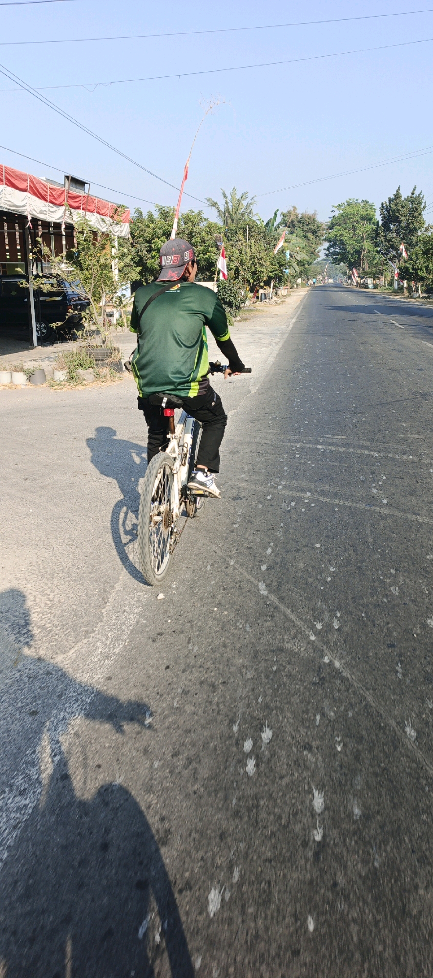 In Mlokorejo, Indonesia, a person is riding a white bicycle on an asphalt road. The cyclist is wearing a dark green long-sleeved shirt with a lime green stripe, black pants, and a baseball cap. They are facing away from the camera, with a dark bag slung across their shoulder. The road is bordered by trees and vegetation on the left and right. Several Indonesian flags, characterized by their red and white colors, are visible along the roadside. The sky is clear and blue, indicating fair weather. The cyclist's shadow is cast on the road to their left. A building with a white and red awning is present on the far left of the image. The road surface appears to have scattered white splotches.