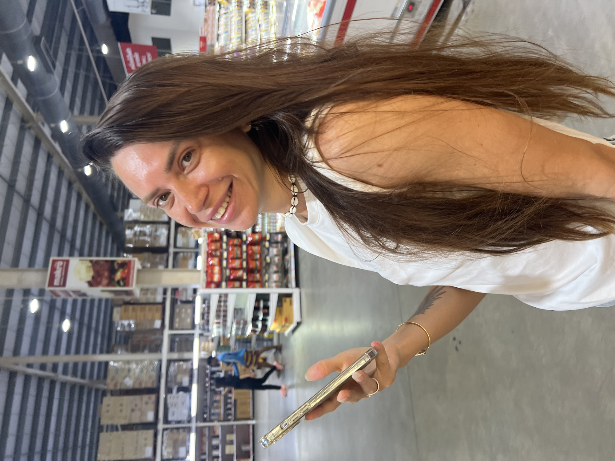 A young woman with long brown hair and a white top is holding a smartphone in her left hand. She is looking at the camera and smiling. She is wearing a seashell necklace and a gold bracelet. She has a tattoo on her left forearm. The background appears to be a large warehouse store, possibly a Makro, as indicated by a sign visible in the upper left portion of the image. Shelves filled with products are visible in the background. The lighting suggests an indoor environment with artificial lights. The floor is concrete. Two individuals are visible in the middle ground, walking away from the camera. The scene is located in Baan Bo Phut, Thailand.