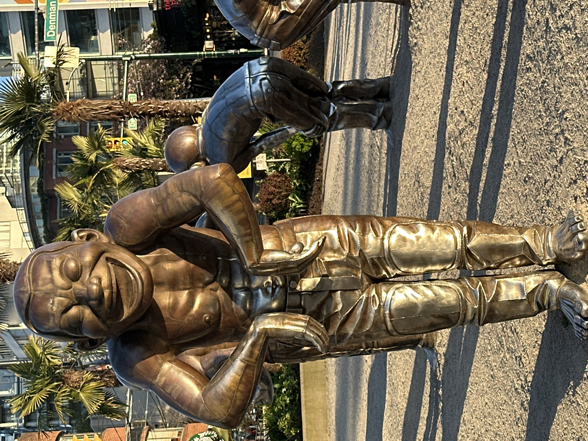The image, oriented vertically, features a large, bronze-colored sculpture of a man depicted with an exaggerated, cartoonish laughing expression. His eyes are closed, and his mouth is wide open, revealing teeth and tongue, conveying intense mirth. His body, segmented and metallic-looking, is positioned as if mid-chuckle, with arms bent at the elbows and hands close to his abdomen.
Behind this central figure, another similar statue is partially visible, bent over at the waist. Both sculptures rest on a light-colored paved surface, which shows long, distinct shadows cast by the figures, indicating bright, direct sunlight, likely from a low angle in the sky (either morning or late afternoon).
The background reveals an urban setting with modern buildings featuring glass windows. Several palm trees are scattered throughout the scene, adding a distinctive characteristic to the environment. A green street sign is clearly visible in the upper left, bearing the text "Denman." A traffic light can also be seen further back among the trees. The overall scene captures public art in a sunny, city environment, consistent with Vancouver, Canada.