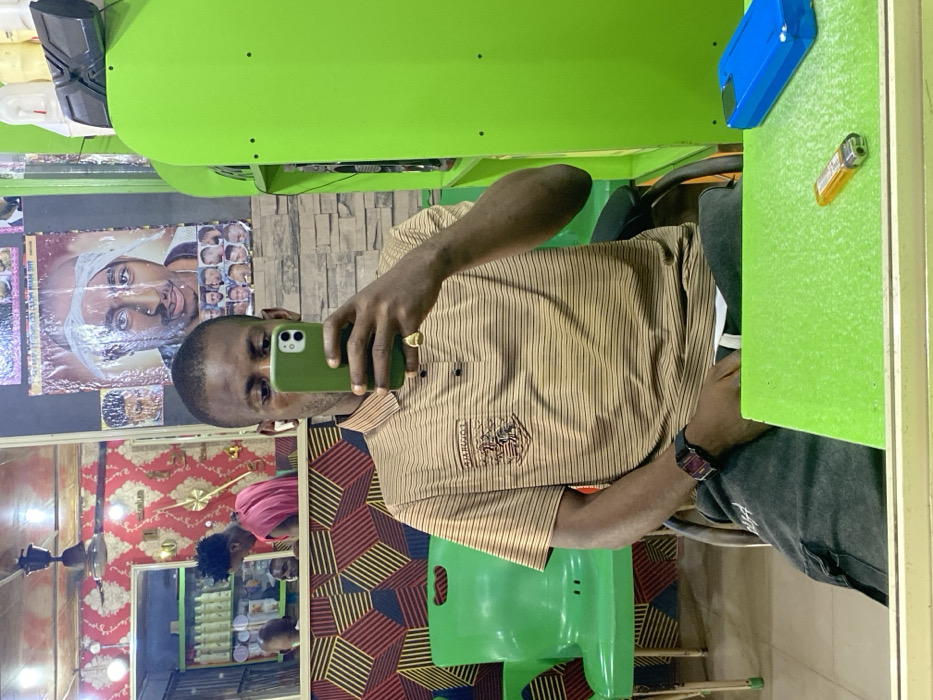 A Black male, wearing a horizontal striped brown and black polo shirt with an emblem, dark grey pants, a black digital watch, and a gold ring on his right hand, takes a mirror selfie in a barbershop or salon located in Zaki Biyem, Nigeria. He holds a dark green cased iPhone, largely obscuring his face, and wears white wireless earbuds. He is seated at a light green counter or table, on which an orange disposable lighter and a bright blue smartphone case are present. The background, visible in the mirror's reflection, includes patterned wallpaper (red and gold, and a geometric red/black/brown design), a stone-textured wall, and posters featuring Tupac Shakur and various hairstyles. In the reflection, another Black male in a red sleeveless shirt is seen attending to a customer in a barber's chair. Additional green plastic chairs, shelves stocked with white product bottles, and a ceiling fan are also visible in the reflection. A solid lime green partition or cabinet section is visible to the subject's right.