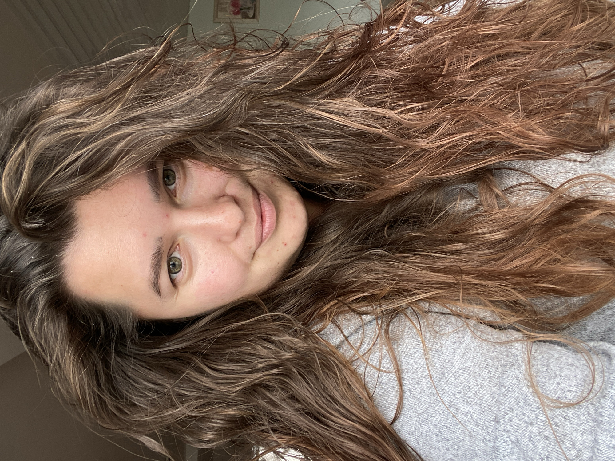 A close-up selfie of a young woman with medium-length, wavy, brown hair is taken indoors. She is wearing a grey sweater. Her face is turned slightly to the right, with her eyes looking directly at the camera. Her hair is loose and falls around her face and shoulders. The background is slightly out of focus, showing what appears to be a wall and a framed picture. There is no indication that this scene is located in the city of Perm, Russia.