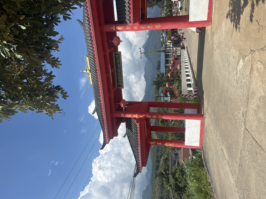The image depicts a red Chinese-style ceremonial gate, characteristic of a Paifang, situated at the entrance to a village identified by a sign on its main horizontal beam as "บ้านสันติสุข" (Ban Santisuk) within "อ.สะเมิง" (Samoeng District), Chiang Mai, Thailand. This location is within or associated with the broader area of Ban Mai Saha Sampan, Thailand.
The gate features a multi-tiered, dark-tiled roof with upturned eaves and is adorned with a golden finial at its apex. Golden Chinese characters are vertically inscribed on the red support pillars, from which red spherical lanterns are suspended. The gate's base is white, contrasting with the vibrant red structure.
A concrete road, exhibiting minor surface cracks, leads through the gate and onto a narrow concrete bridge. The bridge features white railings with patterned square openings, and its initial posts display red and white diagonal striped hazard markings.
Beyond the bridge, the road curves slightly into a village scene. Buildings with predominantly blue or red roofs are visible, some adorned with additional red spherical lanterns. Utility poles with overhead power lines line the road. Two motorcycles are observed moving away from the viewer on the road, indicating vehicular traffic.
The environment is characterized by lush green foliage, including trees and dense vegetation flanking the road and structures. In the background, prominent forested mountains rise under a clear blue sky dotted with large, fluffy white cumulus clouds. Sunlight illuminates the scene, casting shadows, suggesting midday conditions. No specific individuals are discernible in close detail.