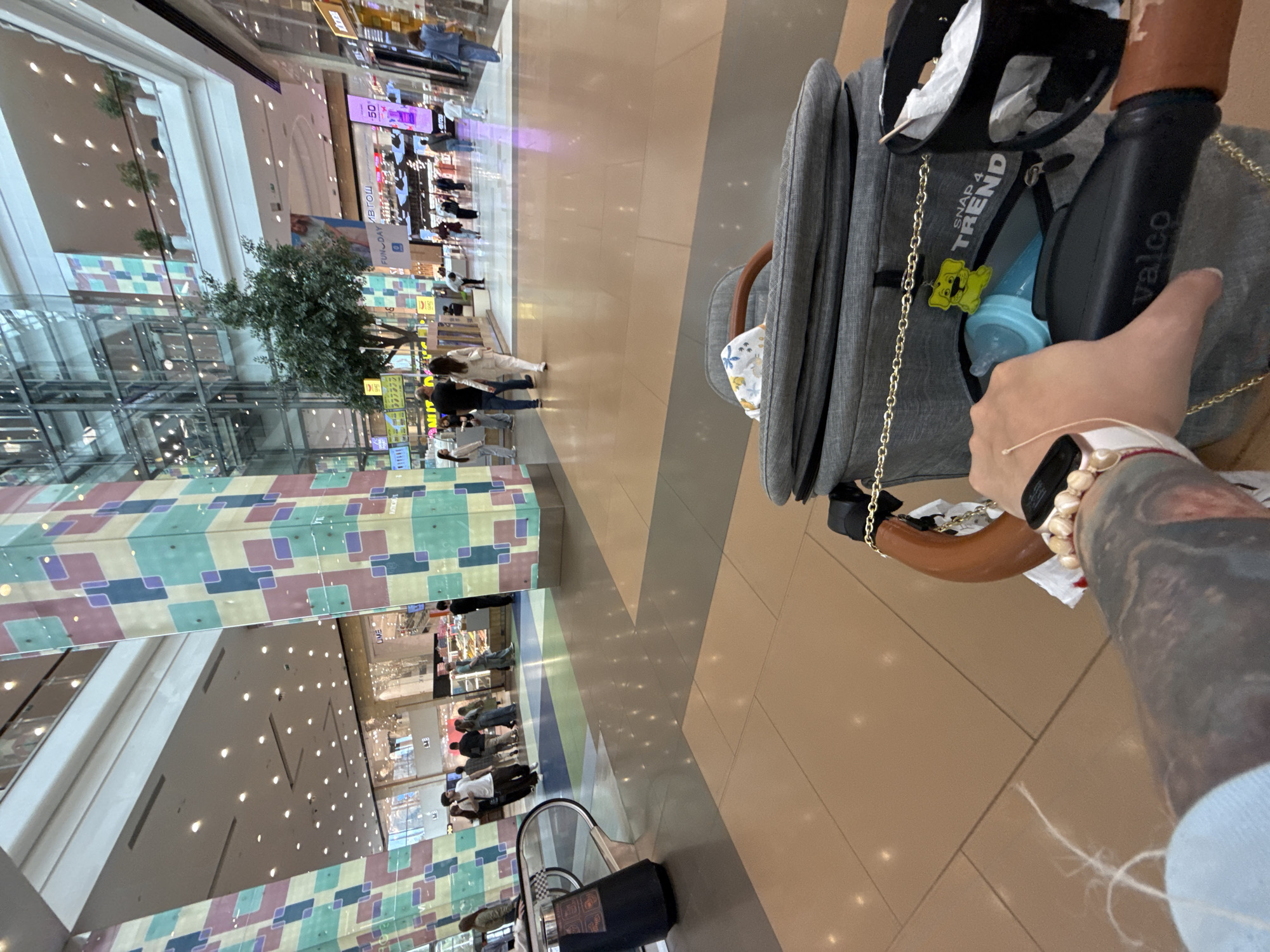 The image shows a first-person perspective of someone in a shopping mall, holding onto the handle of a stroller. The stroller is gray and has a wooden handle. A black water bottle with the brand name "valco" is attached to the stroller. Inside the stroller, a blue baby bottle is visible, along with a yellow charm of a cartoon bear.
The mall's interior is brightly lit with overhead lights. The floor is a neutral-toned tile. In the background, people are walking and shopping. Large colorful panels with geometric patterns adorn a section of the mall's wall. Reflections of the mall's architecture, including its ceilings and lights, are visible in the reflective surfaces. A tree is planted inside the mall, adding a touch of greenery to the environment.
The scene appears to be during the day, as indicated by the bright lighting. There are no clear indicators of the weather outside, but the indoor setting suggests it's not a factor in the immediate scene. The atmosphere seems to be one of everyday activity, with people going about their shopping and daily routines. The image's orientation is somewhat tilted. Text visible includes "valco" on the water bottle and "SNAP 4 TREND" on a pocket of the stroller. The location context is Moscow, Russia.
