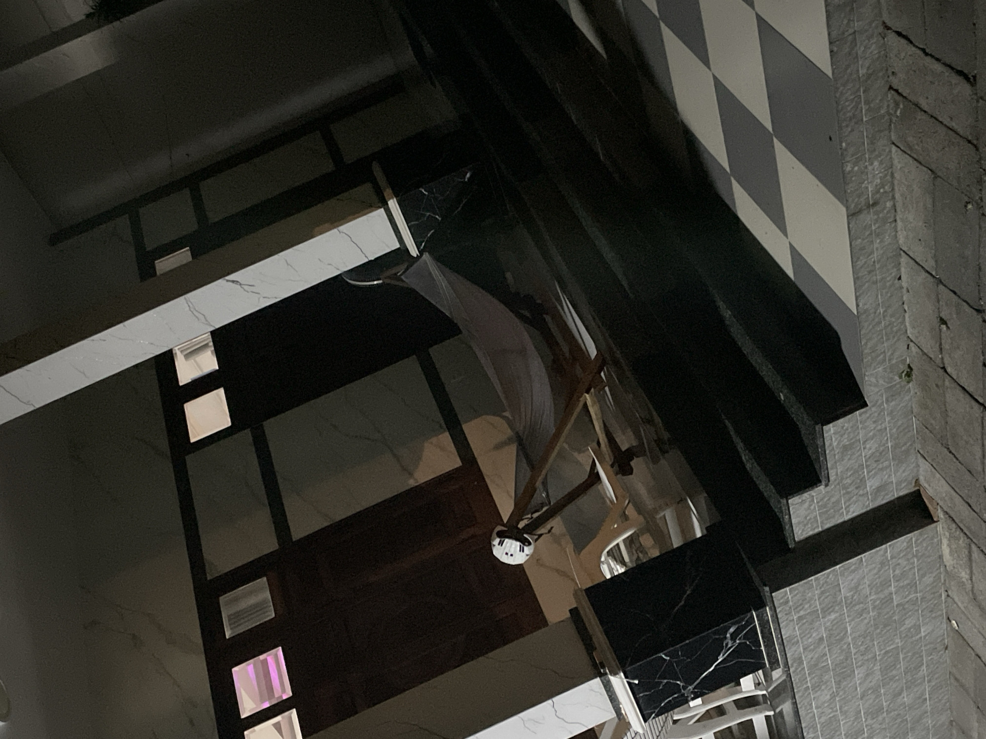 The image depicts a nighttime outdoor scene, likely a patio or walkway area, with a hammock set up in the center. The environment appears to be a residential or possibly a commercial establishment with architectural features such as marble-patterned columns and walls. The ground surface varies, with tiled areas and a checkered pattern visible. A hammock, made of white fabric, is suspended between two points and appears empty. The lighting suggests artificial illumination, casting shadows across the scene. The overall impression is one of a quiet, possibly relaxing setting at night. The location is identified as Phan Thiet Ward, Vietnam, though no specific landmarks or identifying features are visible in the image to further pinpoint the exact location within the ward.
