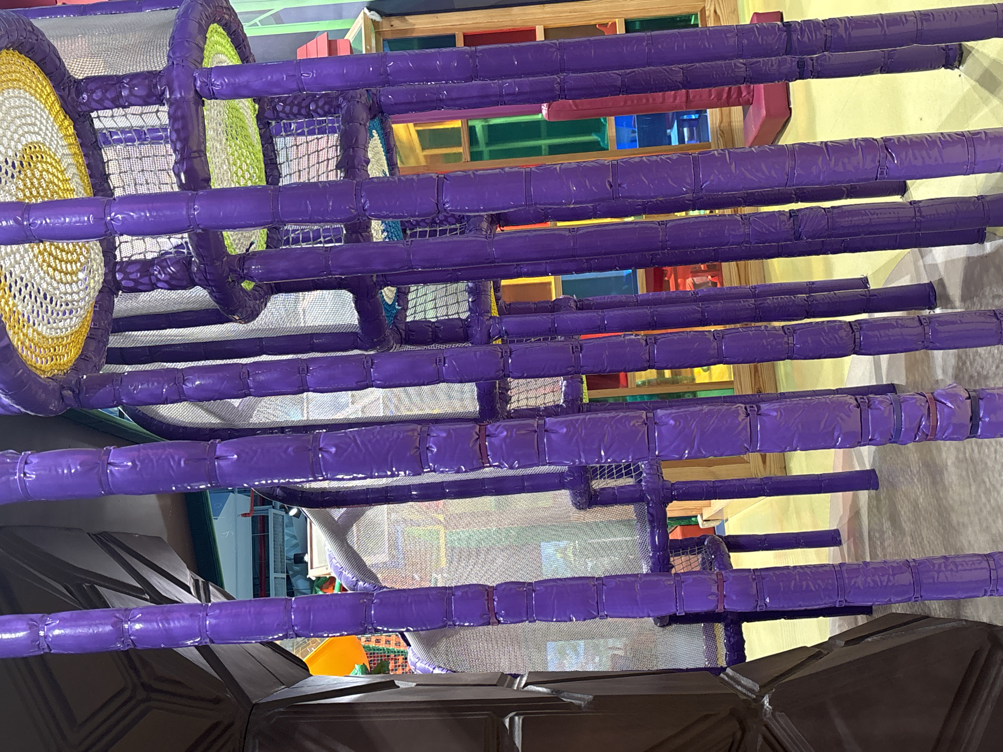 The image depicts the interior of a children's indoor playground in Dubai, United Arab Emirates. The foreground is dominated by tall, cylindrical purple padded poles, some of which are grouped together. These poles are part of a climbing structure with netting visible at the top. Behind the poles, a large, multi-colored structure with wooden framing and translucent colored panels in shades of red, yellow, green, and blue is visible. There are also glimpses of orange and yellow slides. The flooring appears to be a light-colored, soft material. The overall environment suggests a playful and brightly lit space designed for children's recreation.