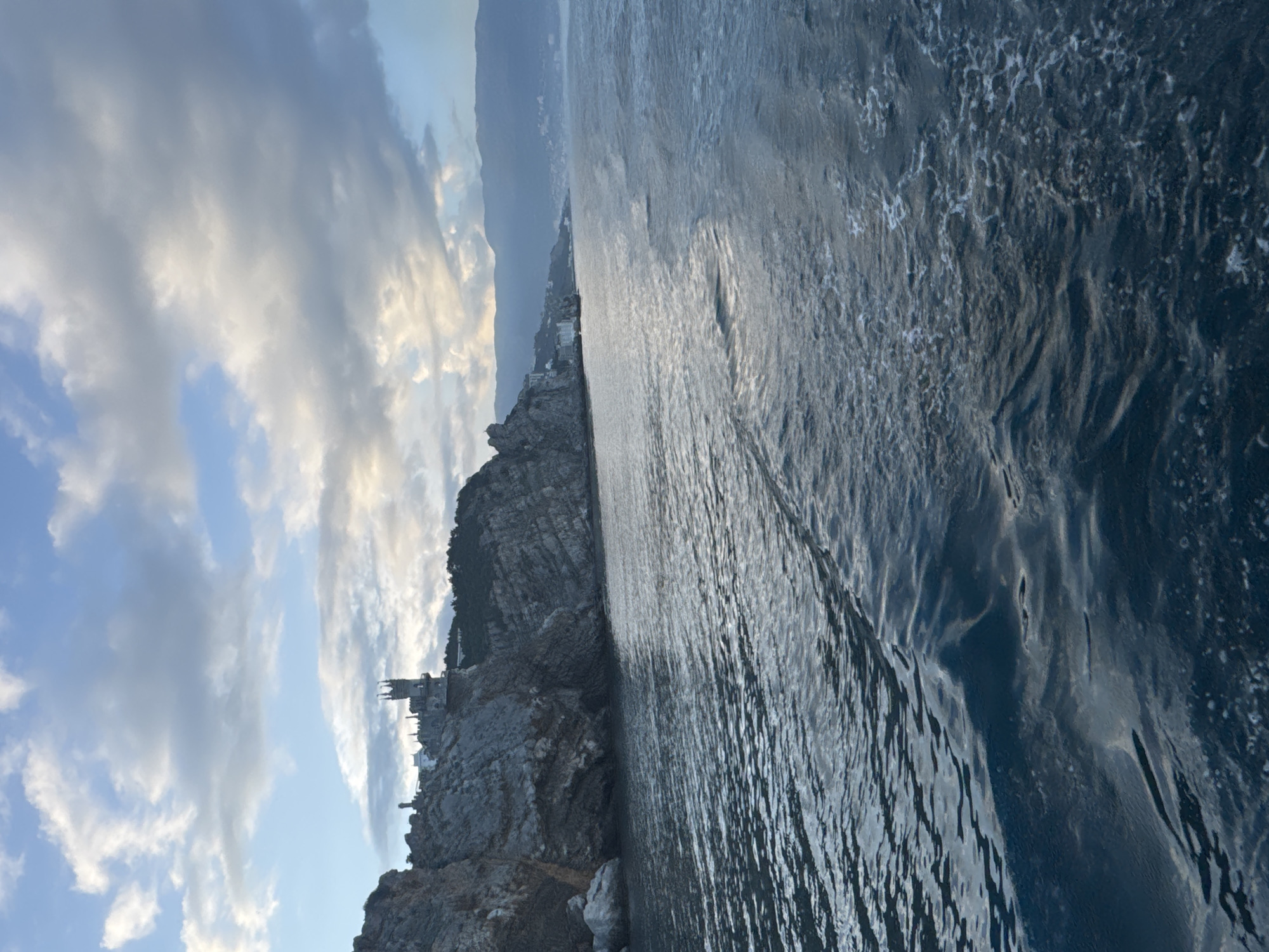 The image shows a coastline with a rocky cliff on the left and the sea on the right. At the edge of the cliff, there is a castle, presumably Swallow's Nest, a popular landmark in Gaspra, Ukraine. The sky above is filled with clouds, with patches of blue visible. The sea appears rough with reflections of the sky. In the background, there is a hazy mountainous landscape. The photograph appears to be taken from a slightly elevated position, possibly from a boat or another cliff overlooking the scene.