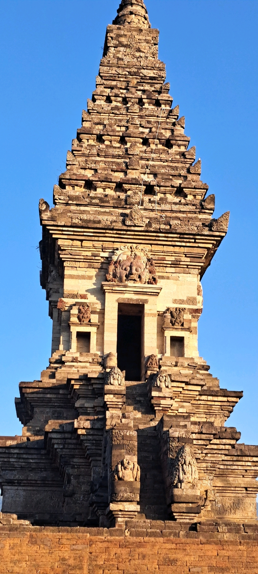 The image is a close-up, vertical, eye-level shot of a multi-tiered, ancient stone temple structure against a clear, bright blue sky. The temple, likely in Candi Wates, Indonesia, is constructed from weathered, reddish-brown bricks and carved stone. Its architecture features stepped levels adorned with intricate carvings of faces and possibly mythological figures. The lower section of the temple has a wide, brick-paved base, leading up to layered stone structures. The main body of the temple exhibits a central entrance with a dark interior, flanked by smaller square openings and decorative carvings. The stepped pinnacle of the temple rises towards the top of the frame, diminishing in size with each level, and is also heavily ornamented with detailed stonework. The lighting suggests it is late afternoon or early morning, casting shadows that highlight the texture and depth of the stonework.