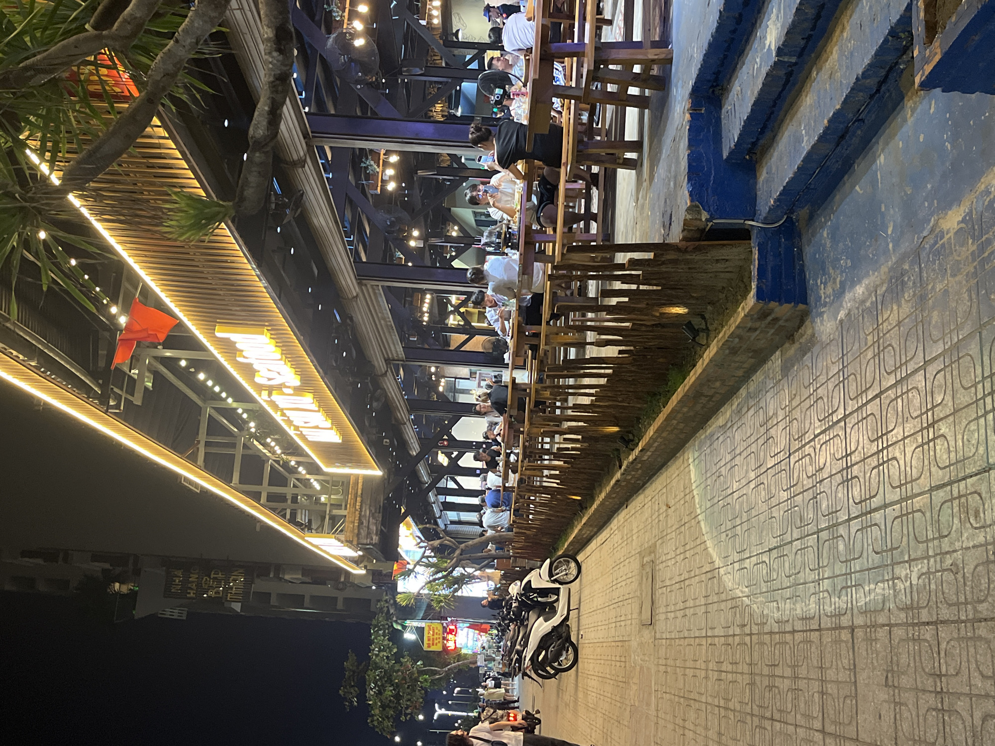 Captured at night in Nha Trang Ward, Vietnam, the image shows a street scene where a restaurant extends over the sidewalk. Patrons are seated at wooden tables on an elevated platform. The restaurant's facade is lit by string lights and bright signage. Parked motorbikes line the sidewalk adjacent to the dining area. The pavement is patterned. People are visible both dining in the restaurant and walking on the street. A tree's branches frame the left side of the view.