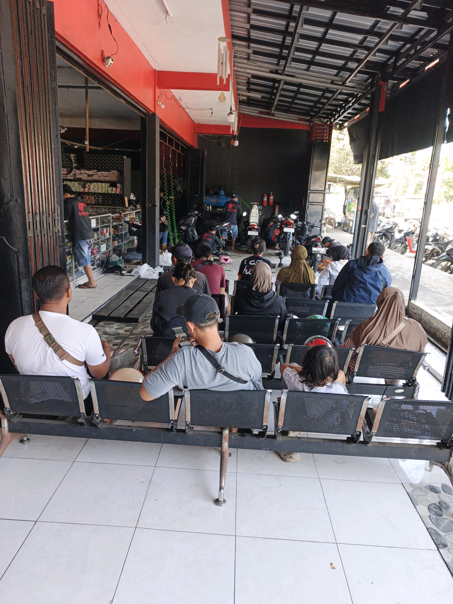 The image depicts an outdoor waiting area in front of a business, likely a motorcycle repair shop or parts store, in Desa Tegalreja, Kecamatan Cilacap Selatan, Indonesia. The facade of the building is painted bright red, with black metal grating and a corrugated metal roof. A variety of motorcycles are parked in the background, some of which are on a lift. Inside the store, shelves stocked with goods are visible through an open doorway.
In the foreground, several people are seated on black metal benches, facing the building. Most are looking towards the entrance or at their phones. The individuals are dressed casually, and several women are wearing hijabs. The ground is tiled with white square tiles. The overall scene suggests a busy day at a local business, with customers waiting for service or to make purchases. The lighting indicates it is daytime, with sunlight casting shadows.