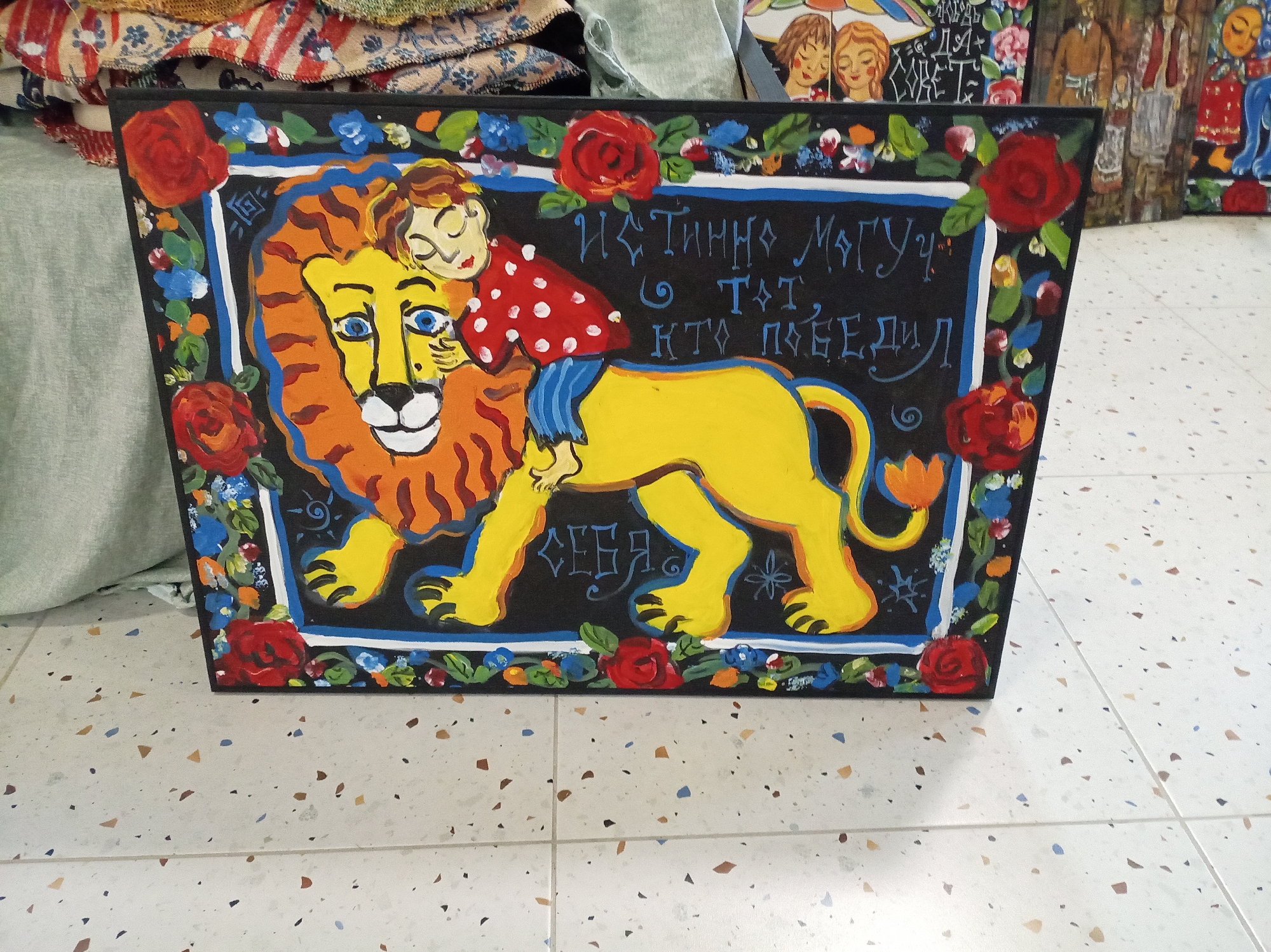 A painted wooden board depicts a lion with a child riding on its back, surrounded by a floral border. The lion has a yellow body and a mane rendered in shades of orange and brown. The child is wearing a red shirt with white polka dots and blue pants. The board is decorated with red roses and smaller blue and white flowers, framing the central image. The text in Ukrainian reads "Істинно магуч той хто переміг себе" (truly mighty is he who conquers himself). The artwork rests on a tile floor with a speckled pattern. The scene is in an unspecified location.