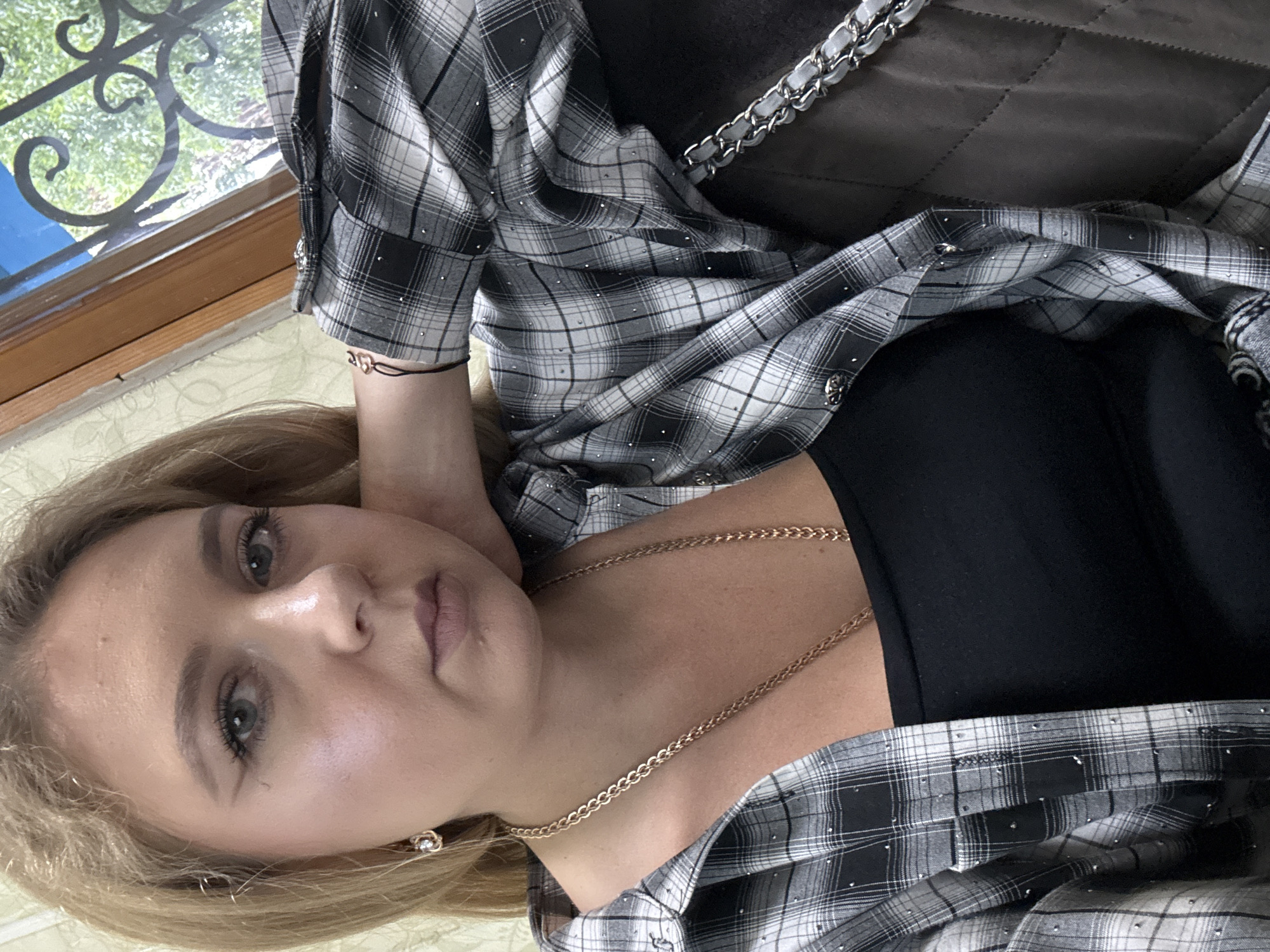 A woman with blonde hair and makeup is pictured from the chest up. She is wearing a black tank top and an open plaid shirt decorated with small rhinestones. She has on a gold necklace and earrings, and a bracelet on her wrist. The woman is leaning her head on her hand, looking towards the camera. She appears to be sitting indoors near a window with a view of trees and greenery. The background wall has a textured pattern.