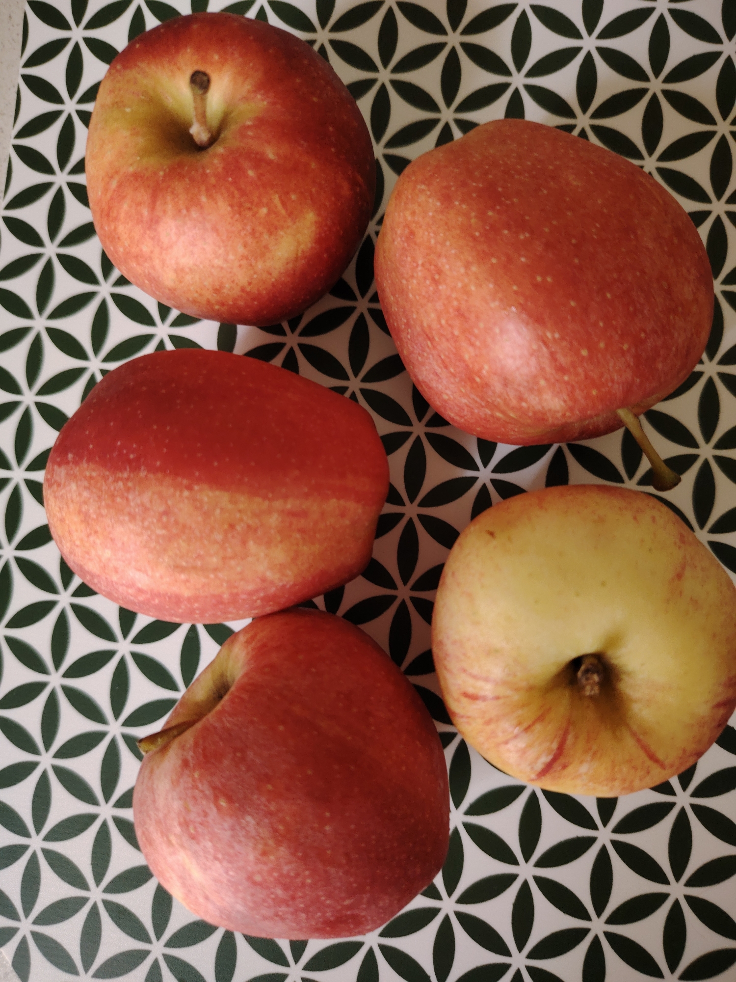 Five red and yellow apples are arranged on a patterned surface. The surface has a repeating design of interlocking green leaf shapes on a white background. The apples vary in ripeness and color, with some exhibiting more red and others more yellow. One apple in the bottom right is cut in half, revealing its pale yellow flesh. There is no indication of a city or location within the image itself, and therefore no information can be provided about Кривец, Russia.
