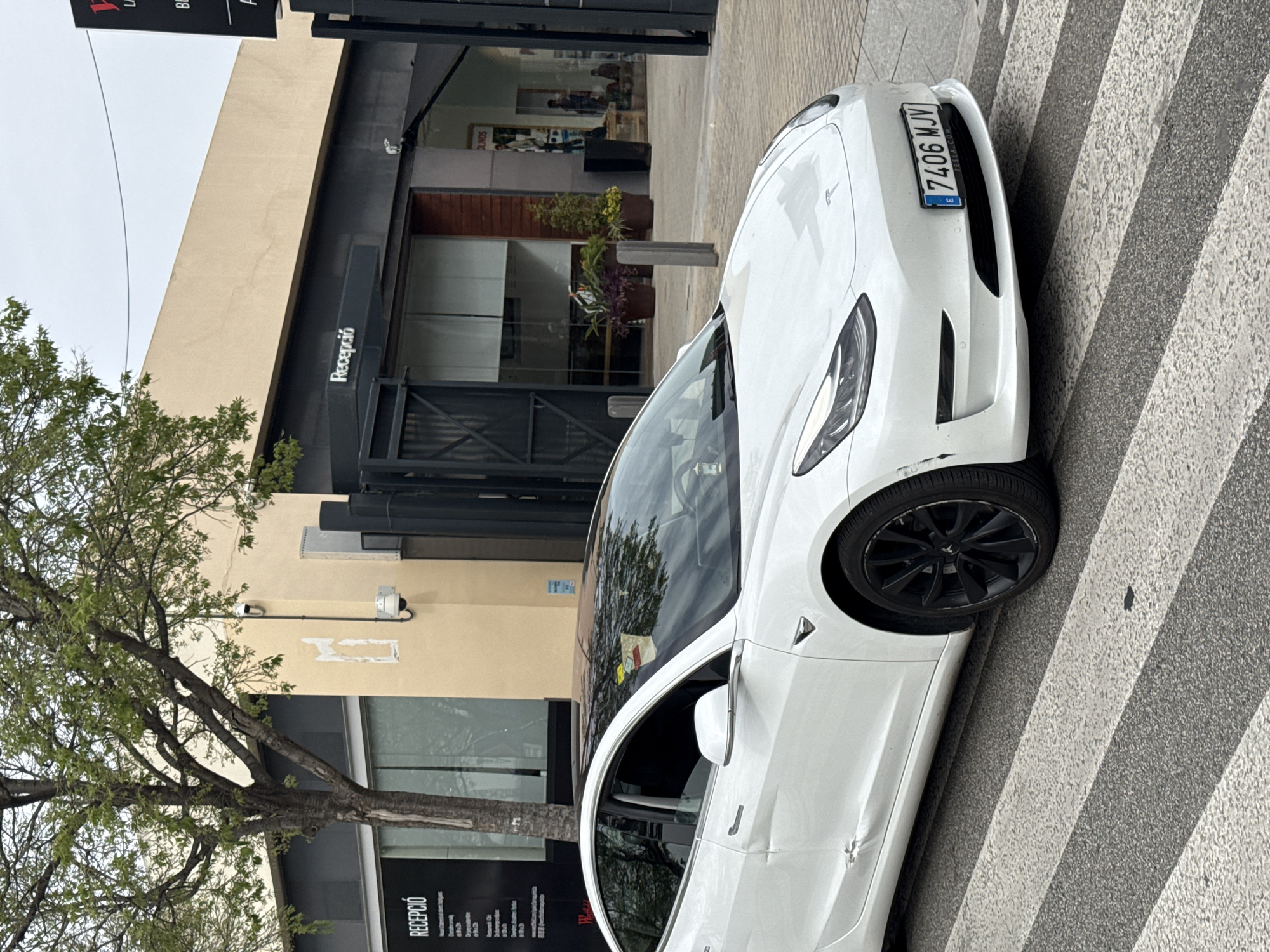 A white Tesla Model 3 is parked on a crosswalk in Barcelona, Spain. The car's license plate reads "7406 MJV". The background features a building with a sign that reads "Recepció" (Reception in Catalan) and a tree on the left. The day appears overcast.