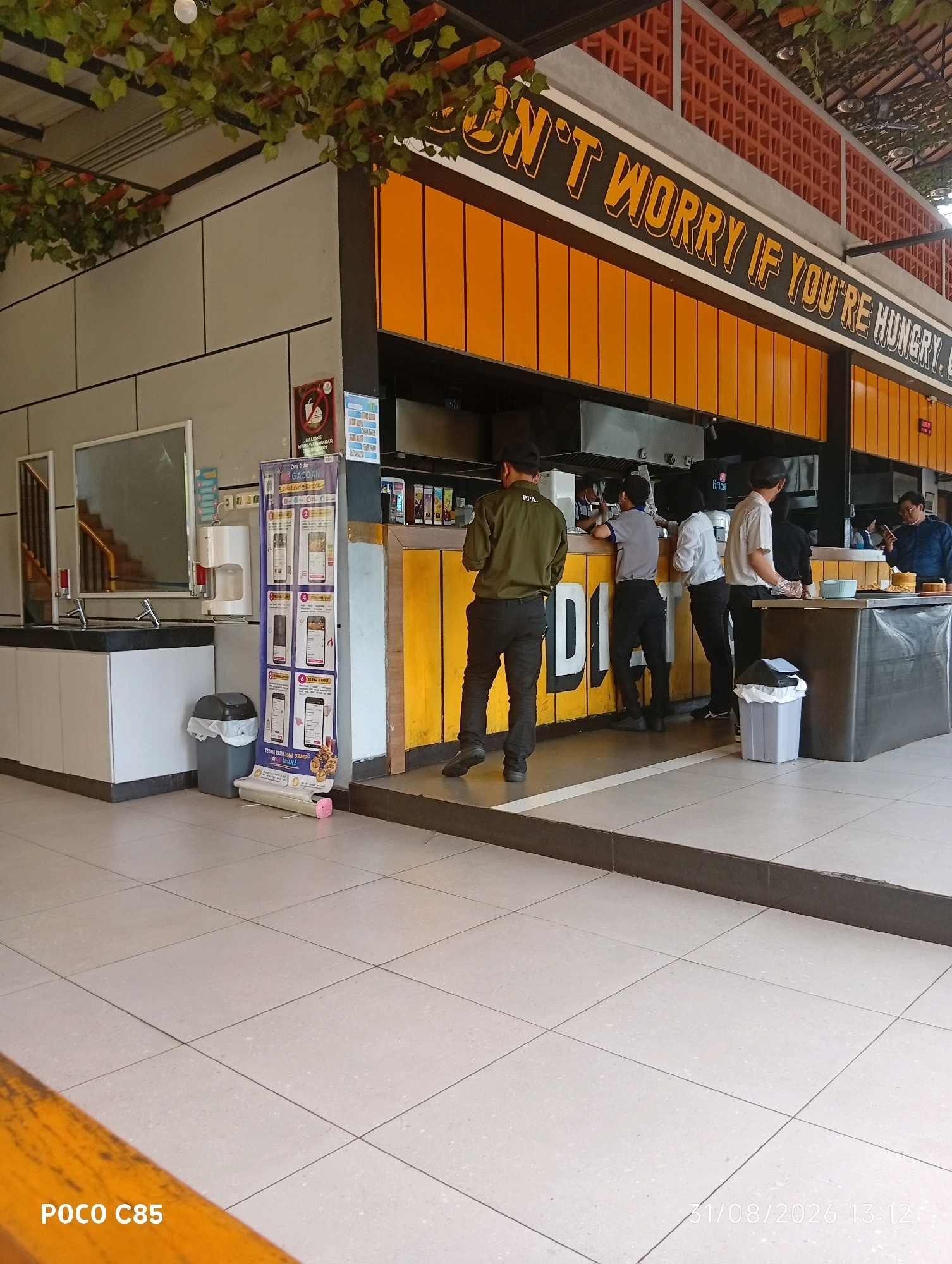 This image depicts an outdoor eating area with a food stall. The stall has a bright orange exterior and a sign that reads "DON'T WORRY IF YOU'RE HUNGRY. LET'S GO." Several people are visible, some appearing to be employees of the stall and others customers. One person in a green shirt is standing with their back to the camera, looking towards the stall. Another person in a grey shirt is leaning against the counter. Behind them, several other individuals are interacting, suggesting a busy service environment. In the foreground, the tiled floor is visible, and a trash can is placed near the stall. The overall impression is of a casual, bustling eatery. The presence of the "POCO C85" watermark and the timestamp "31/08/2026 13:12" suggest this image was taken with a Poco C85 phone on August 31, 2026, at 1:12 PM. Given the context of the Indonesian language on the poster and the likely style of the establishment, this scene is probably located in South Tangerang, Indonesia.